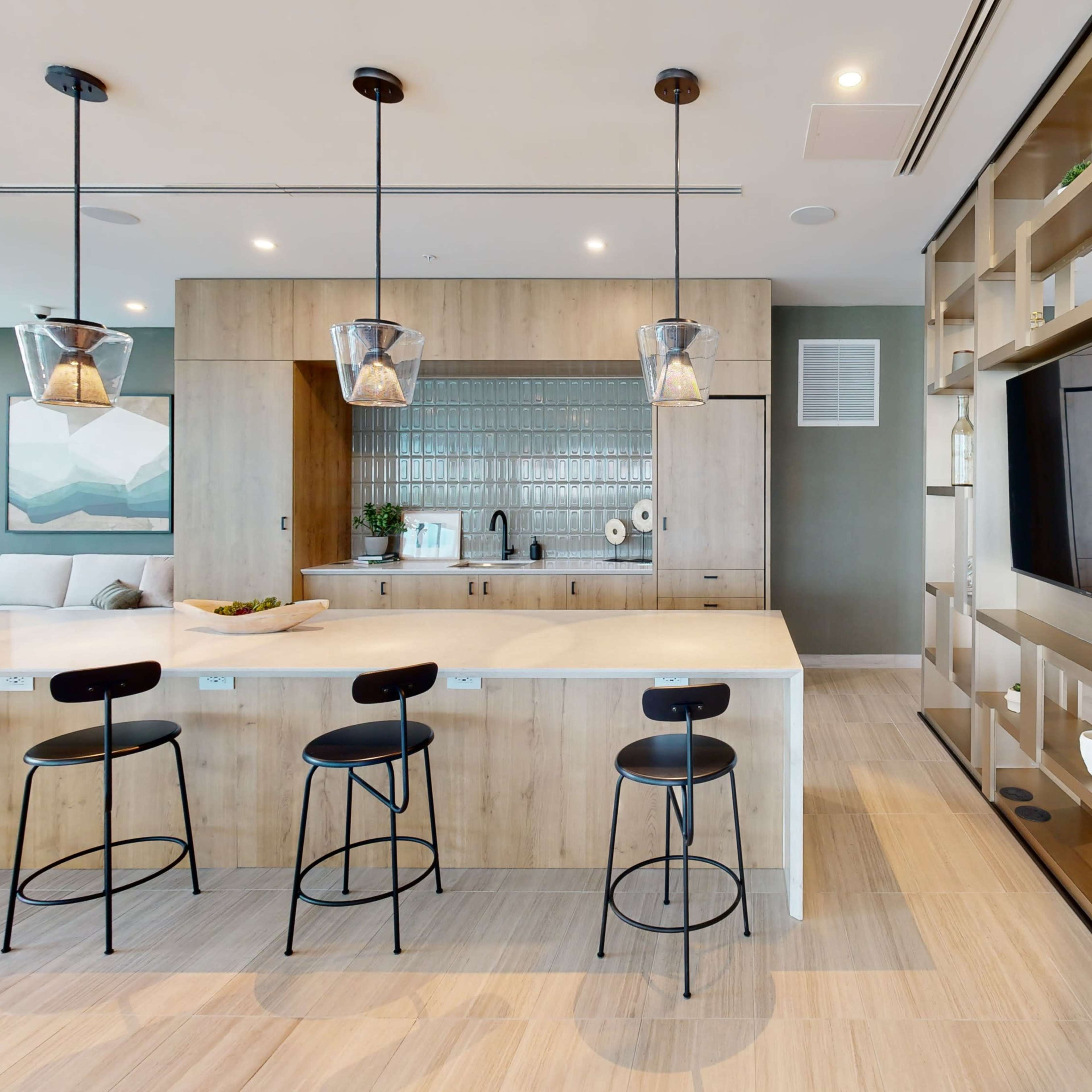A modern kitchen area features a central island with seating, pendant lights, and a wall-mounted television alongside built-in shelving.