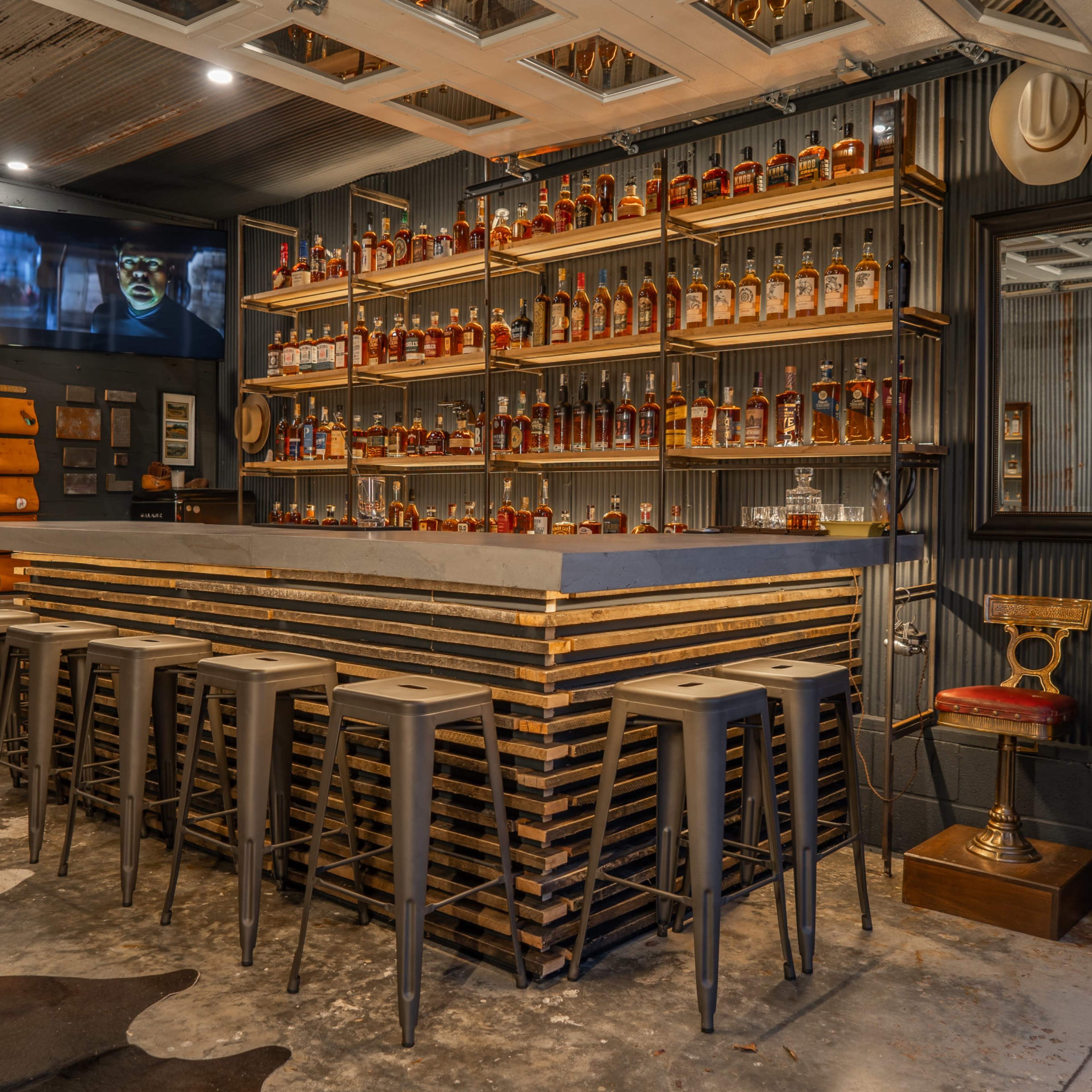 A bar area with a wooden shelf filled with various bottles of whiskey, a concrete bar top, and metal stools arranged in front.