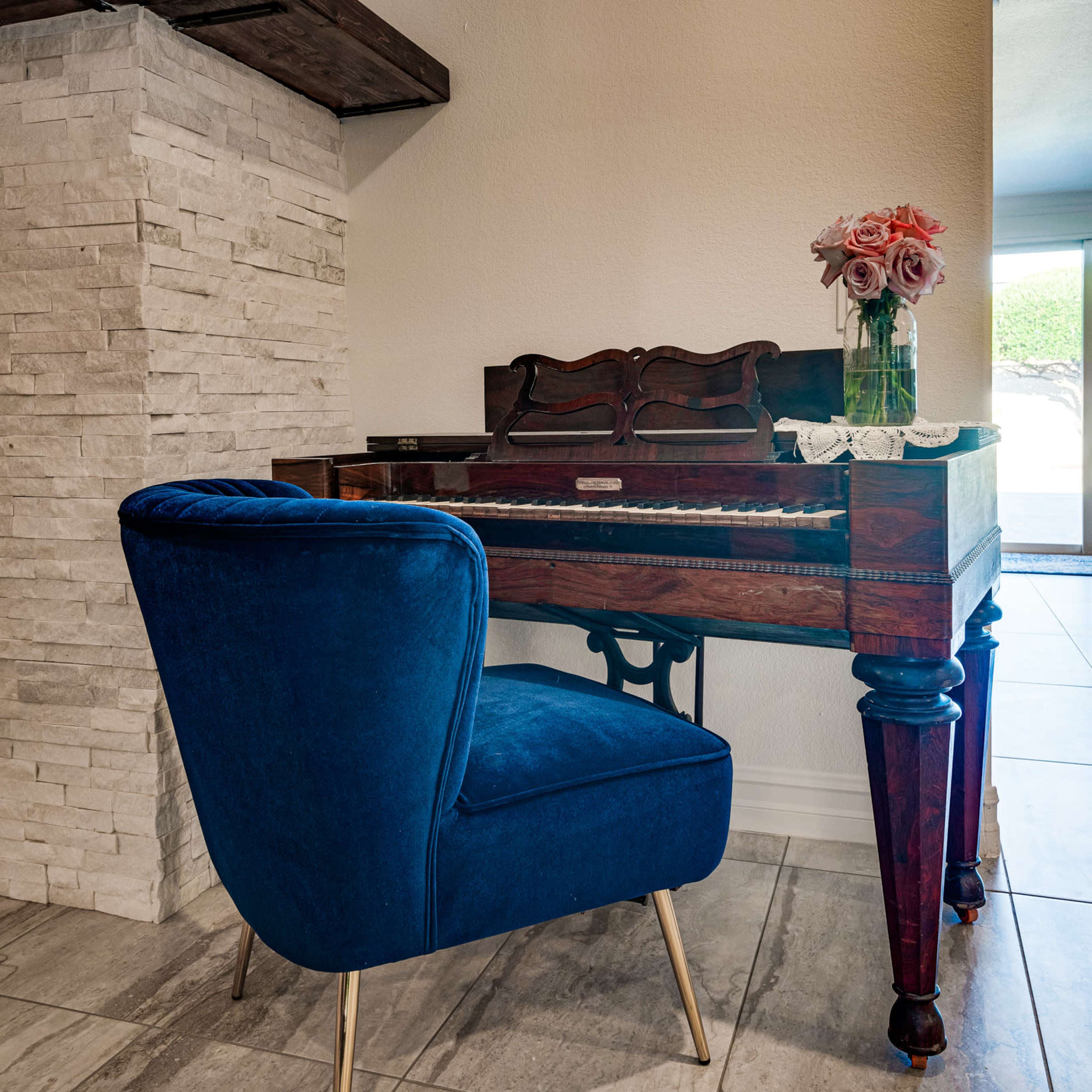 A vintage piano is placed beside a blue velvet chair and a vase of roses on a lace doily, against a stone accent wall.