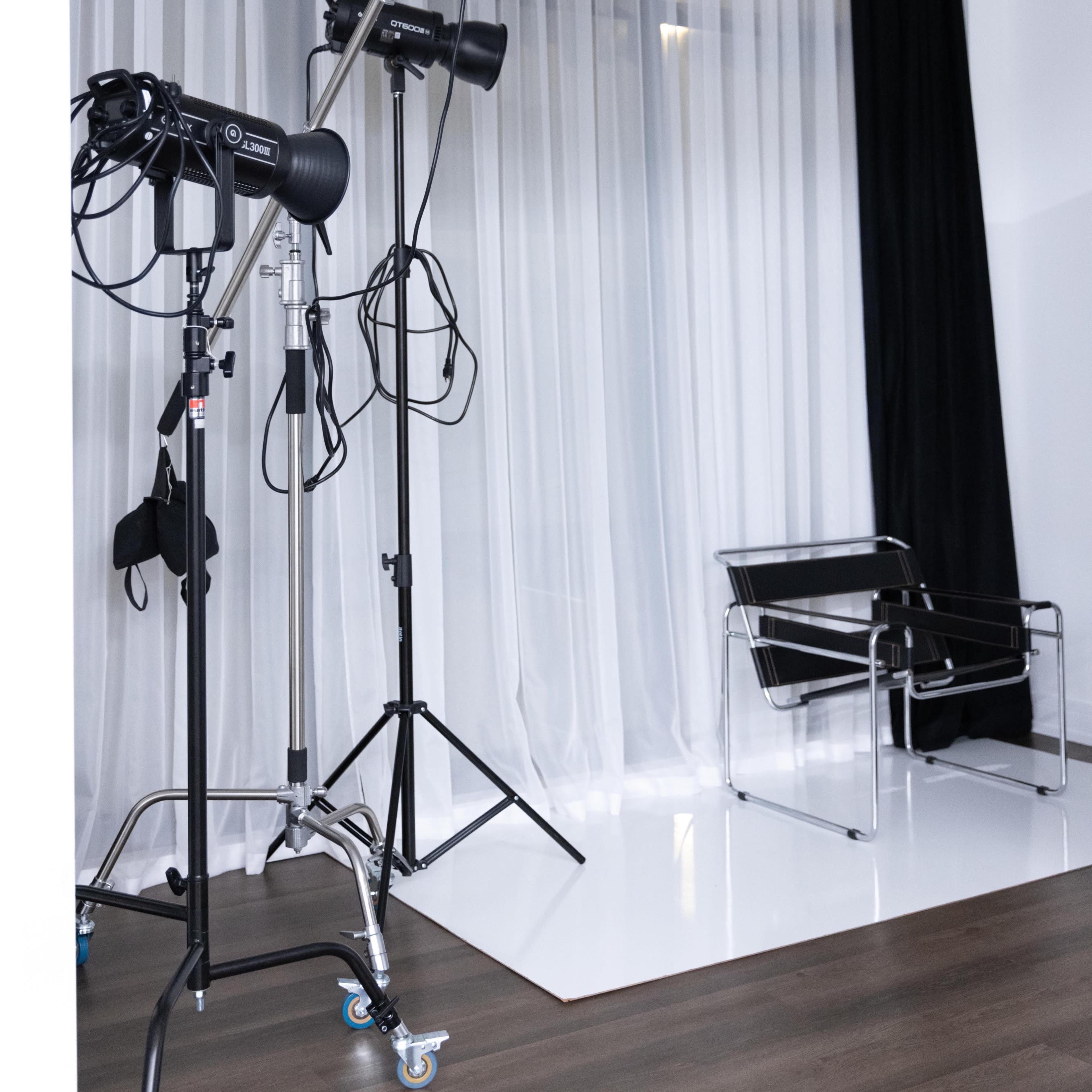 A photography studio setup with softboxes, lighting equipment, and a modern chair against a backdrop of white curtains and dark drapes.