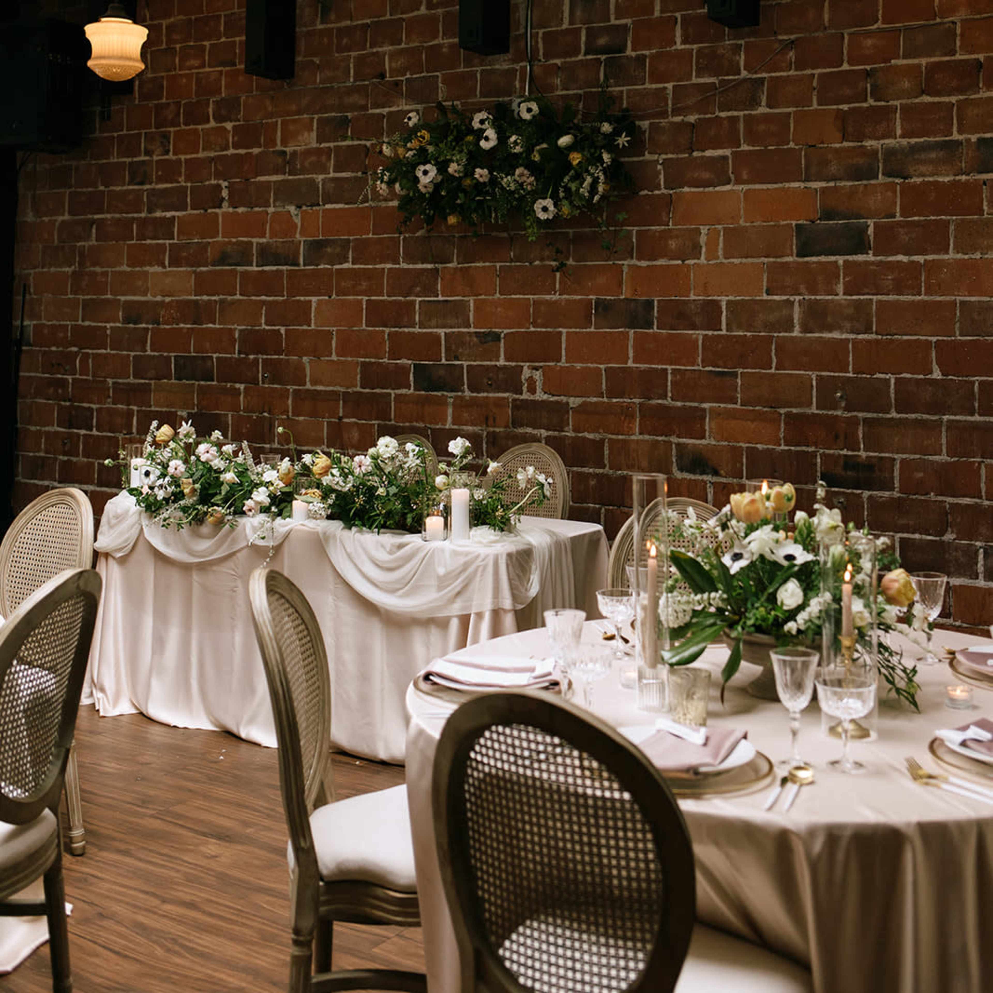 A dining setup with elegantly arranged tables adorned with floral centerpieces and candles, set against a brick wall.