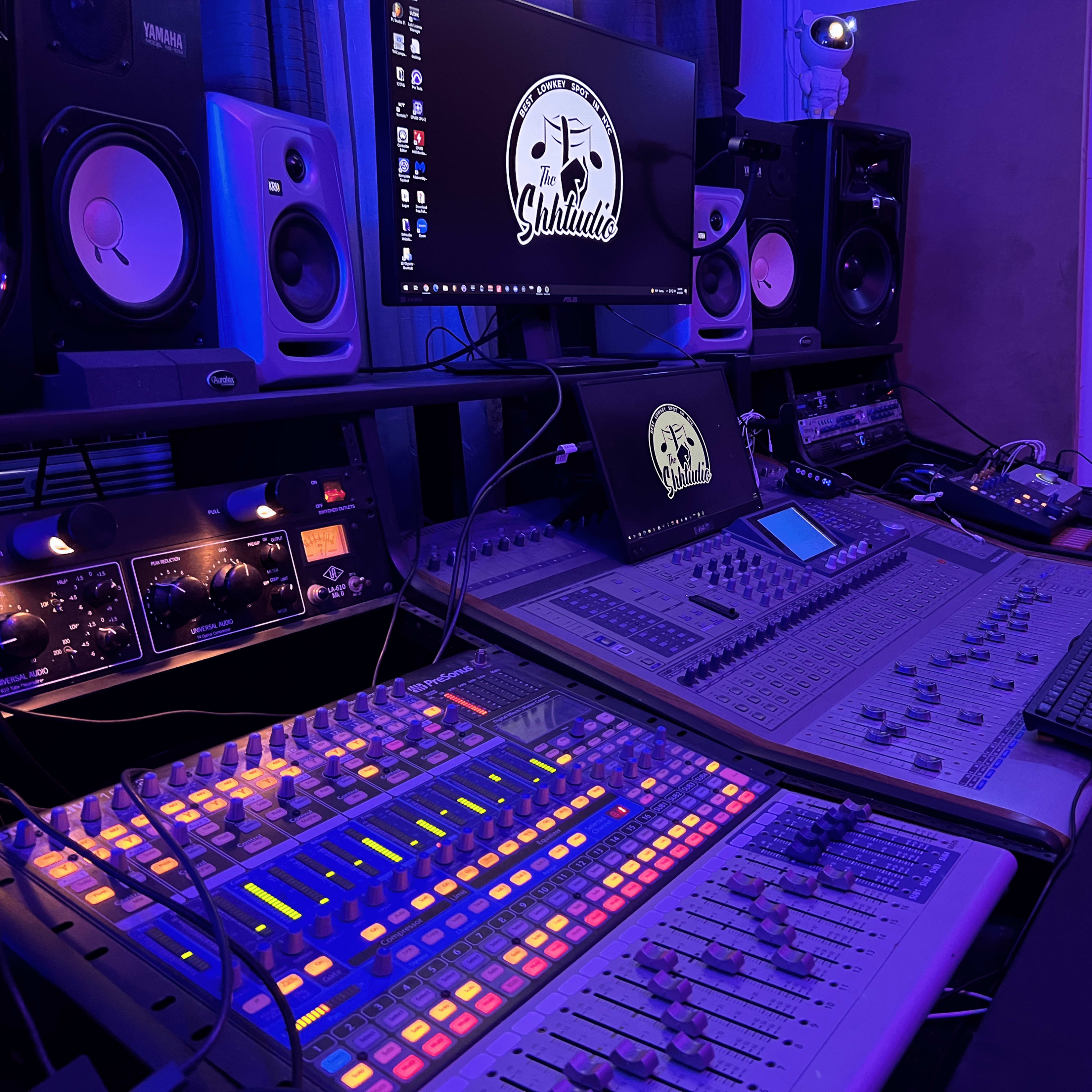 A music production studio with a mixing console, computer monitor, and various audio equipment surrounded by blue lighting.