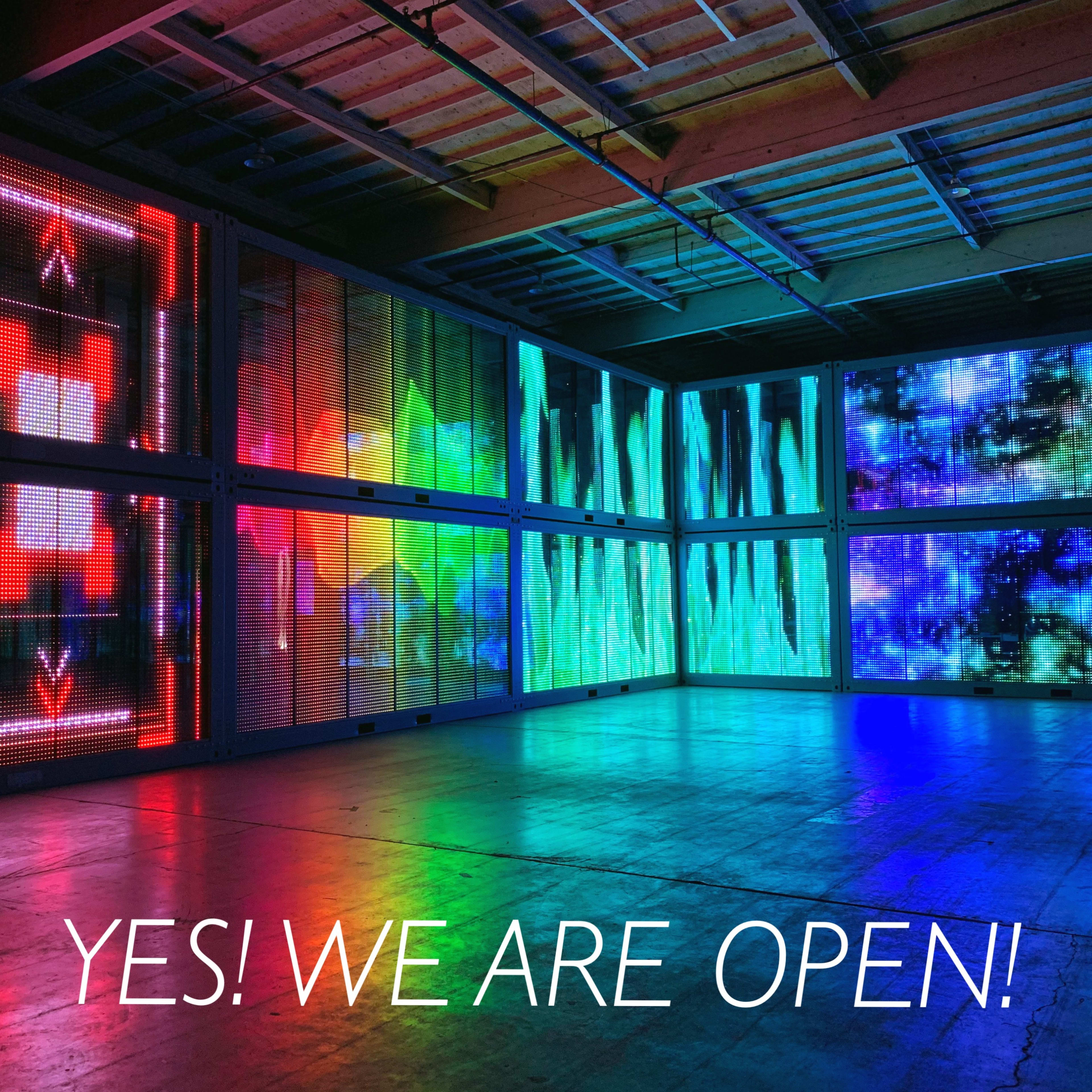 A colorful, illuminated art installation in a spacious room with a sign stating, "YES! WE ARE OPEN!"