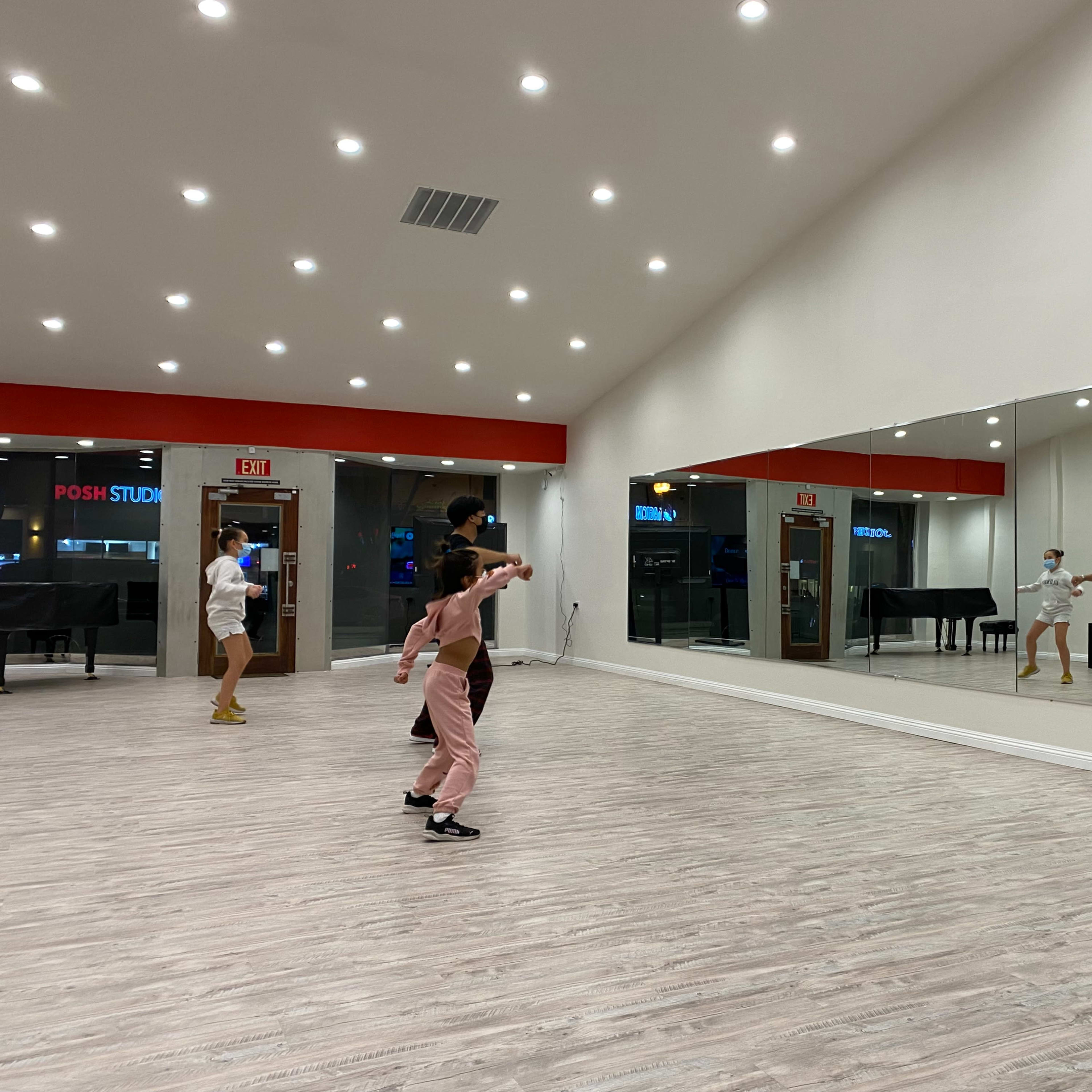 A group of dancers practices in a brightly lit studio with a large mirror and wooden flooring.