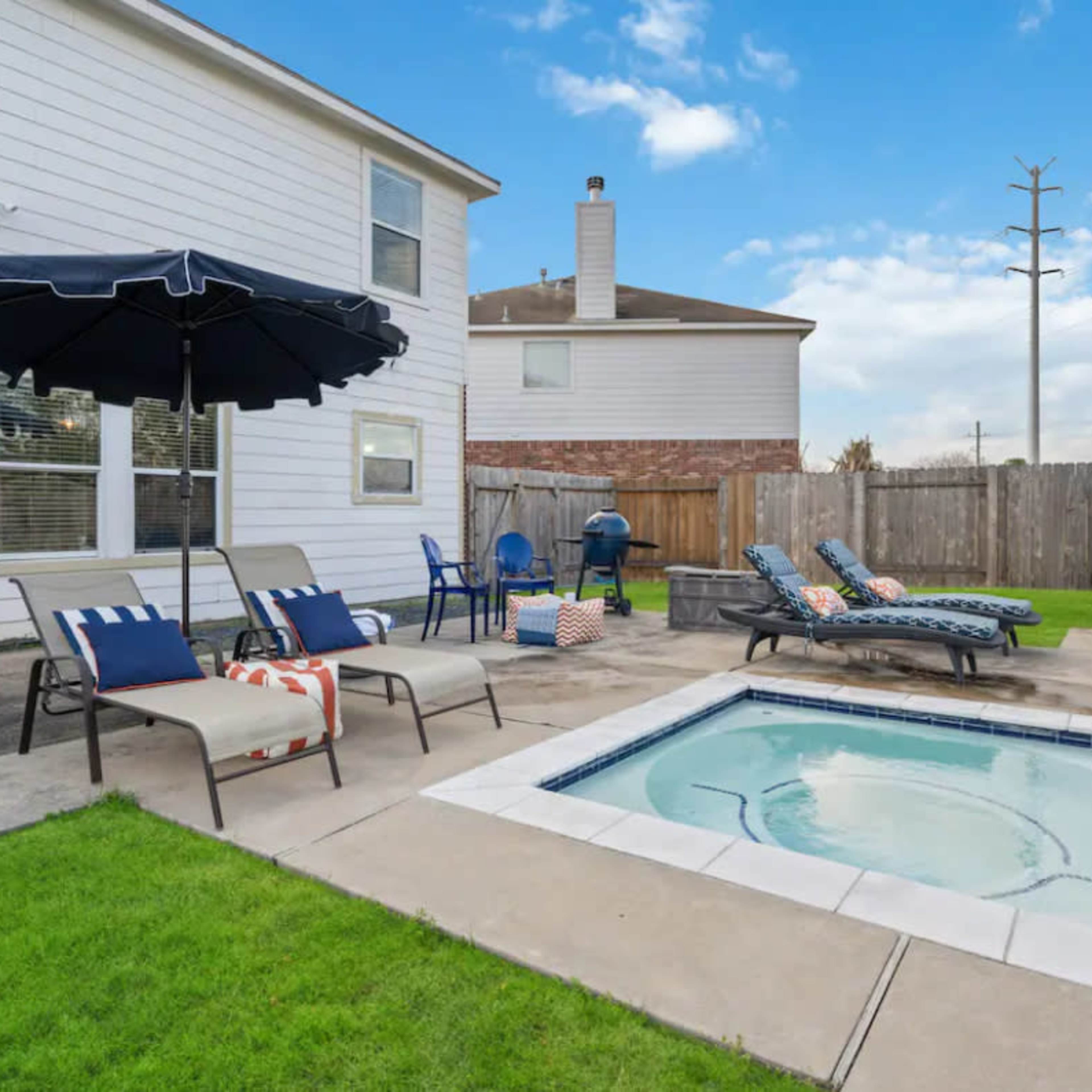 A backyard with a swimming pool, hot tub, lounge chairs, and a patio umbrella beside a two-story house.
