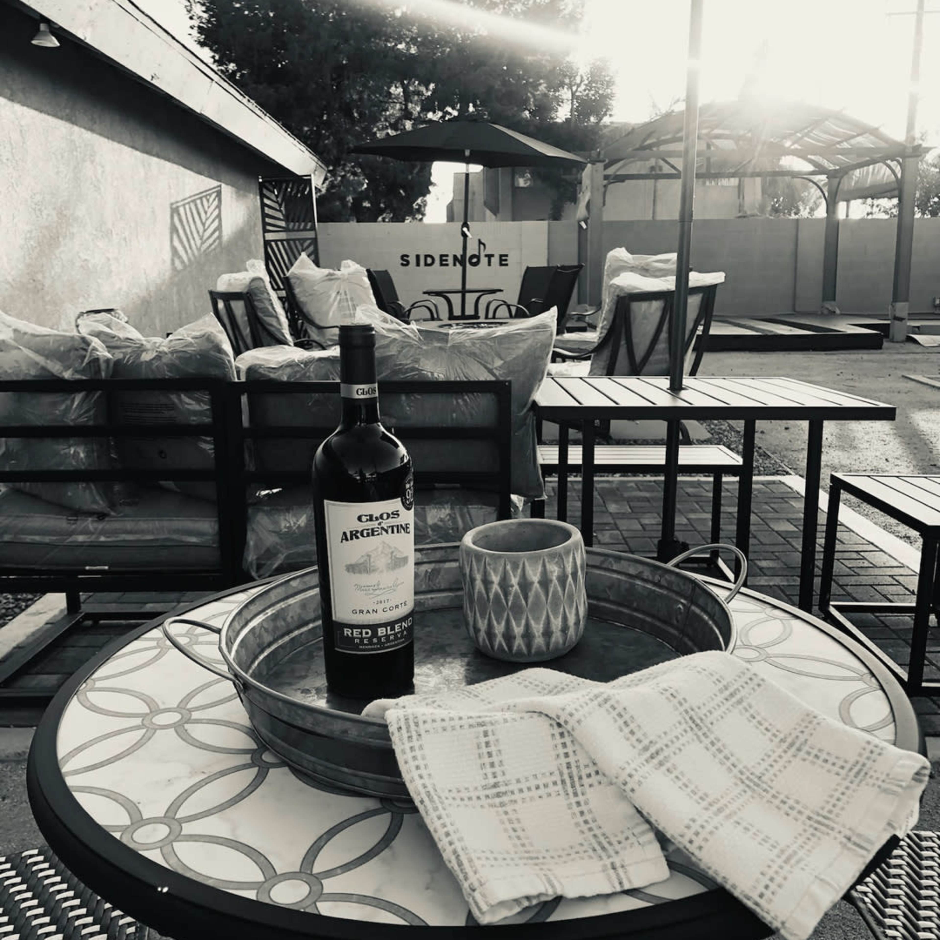A bottle of wine and a mug are placed on a round table, with empty chairs and outdoor seating in the background.