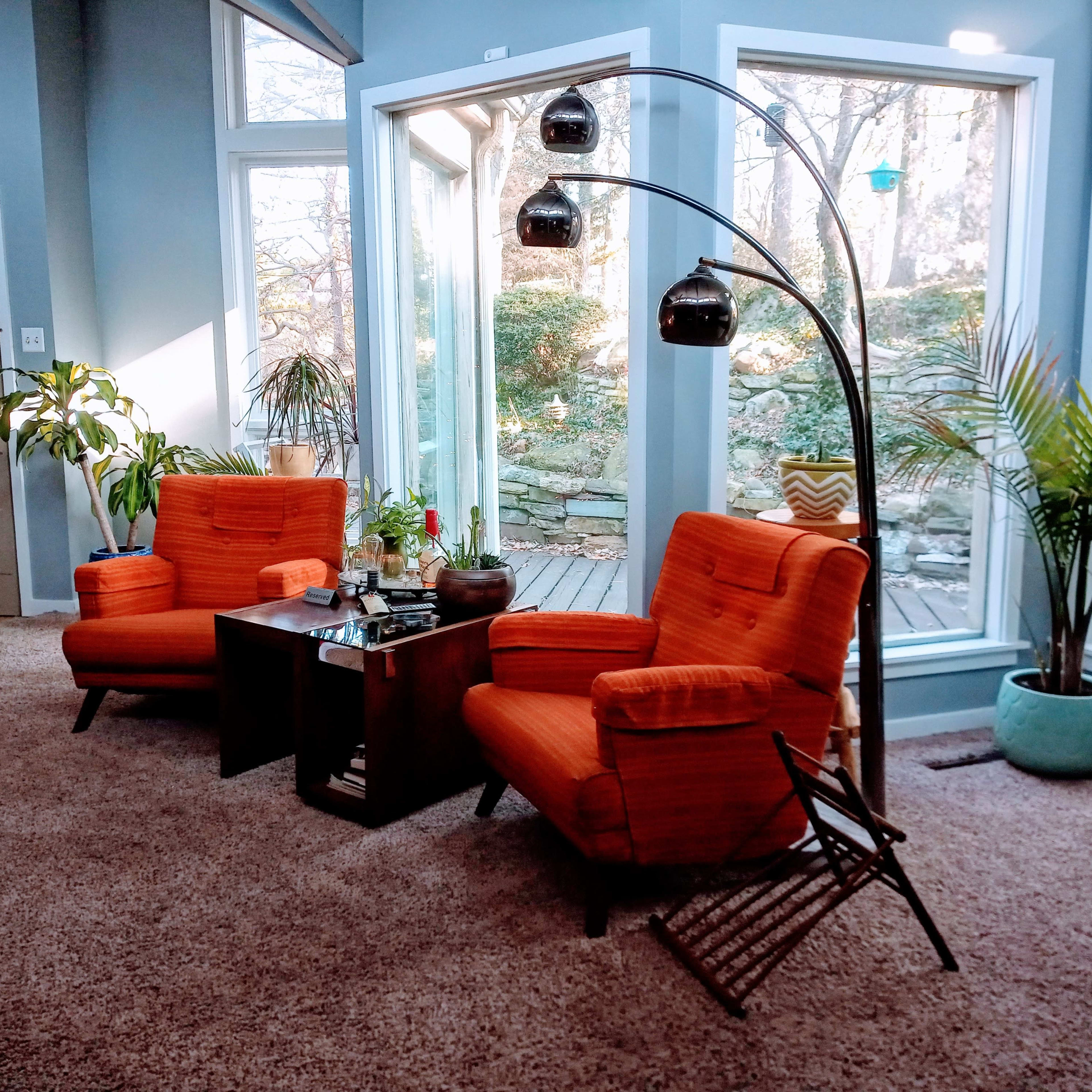 Two orange armchairs positioned in a brightly lit room with large windows and a view of greenery outside.