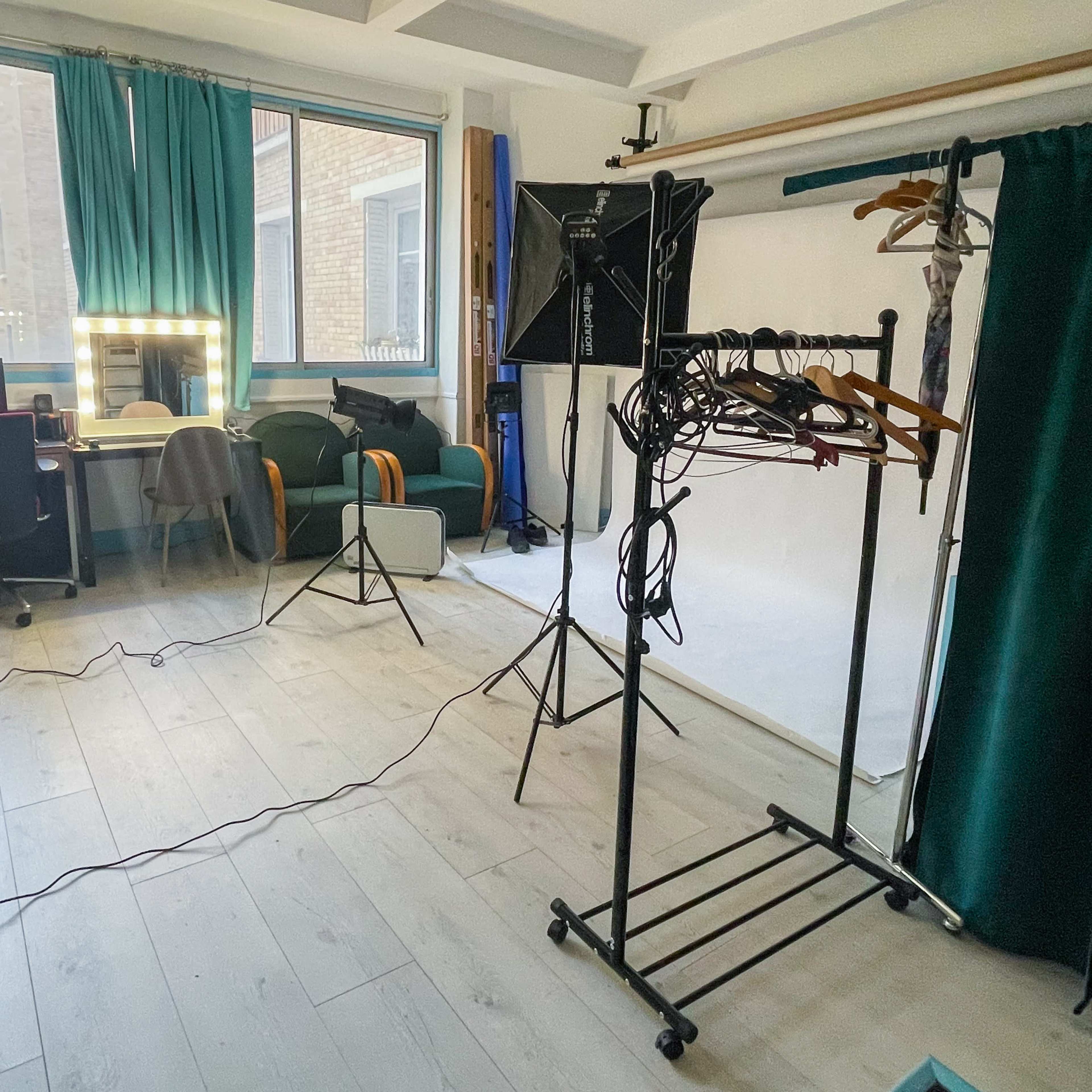 A well-lit studio space with a desk, a mirror, a green chair, a clothing rack, and a backdrop setup.