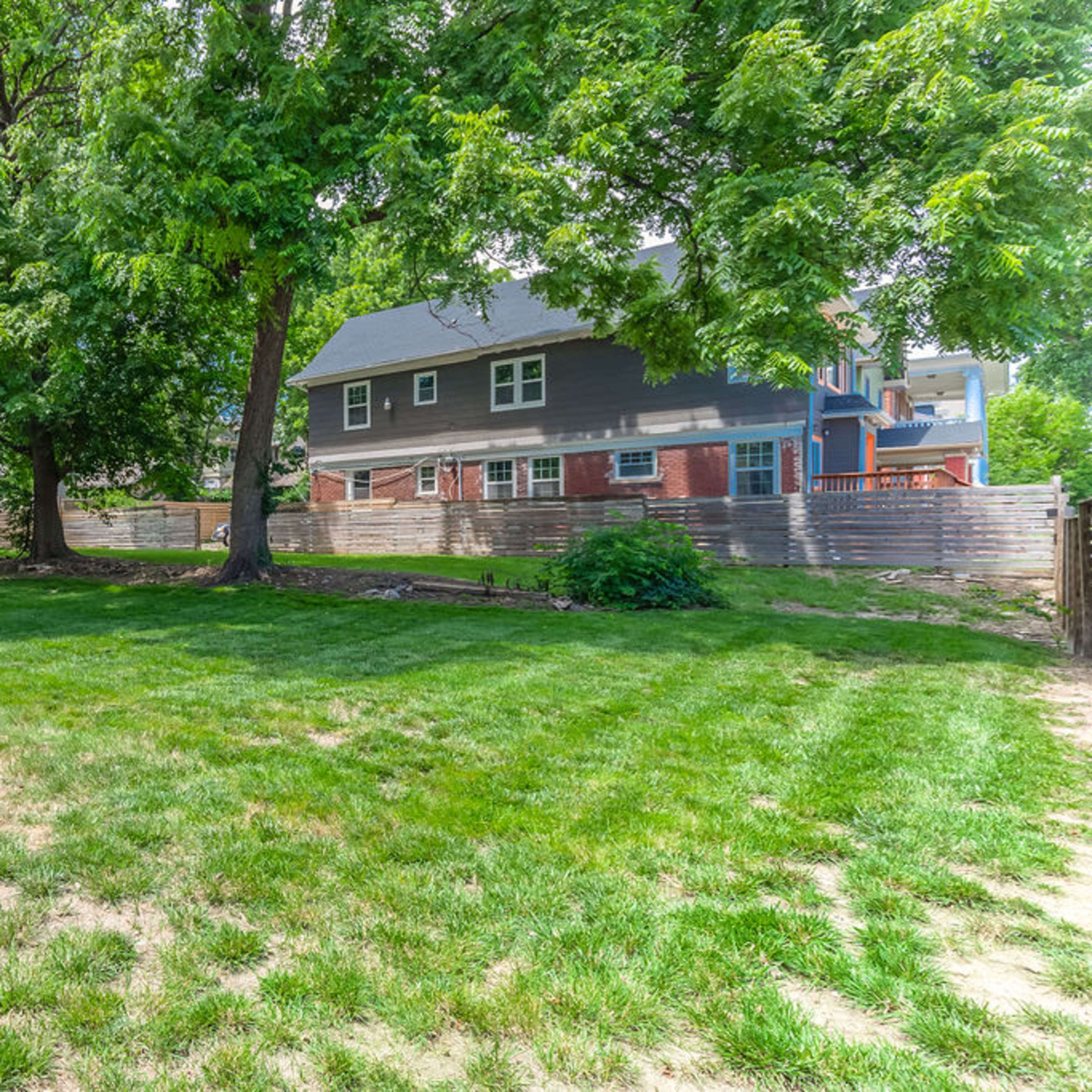 A spacious backyard with a wooden deck, green grass, and several large trees, alongside a two-story house in the background.