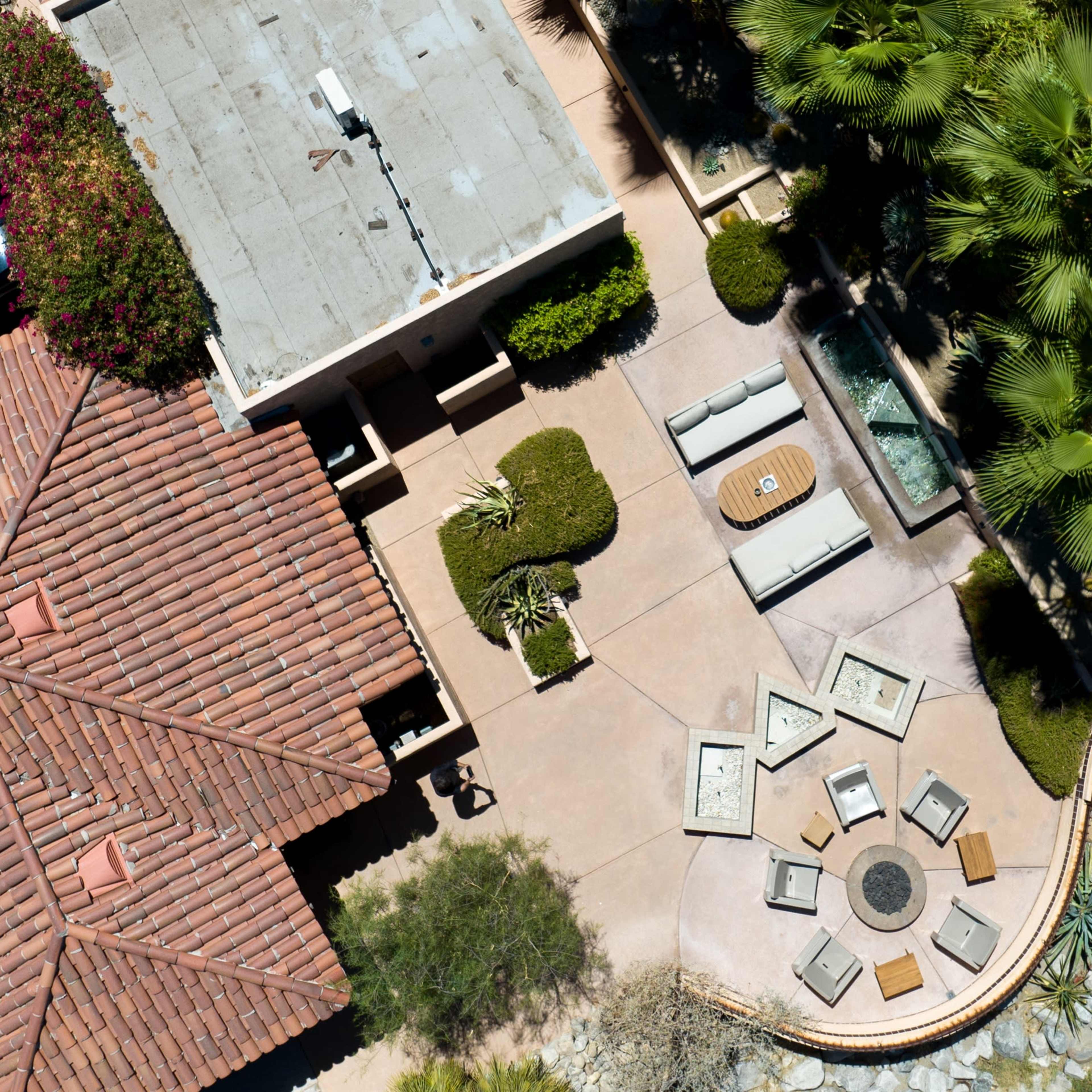 An aerial view shows a modern home with a red tiled roof, a patio featuring circular seating, and landscaped greenery surrounding a pool area.