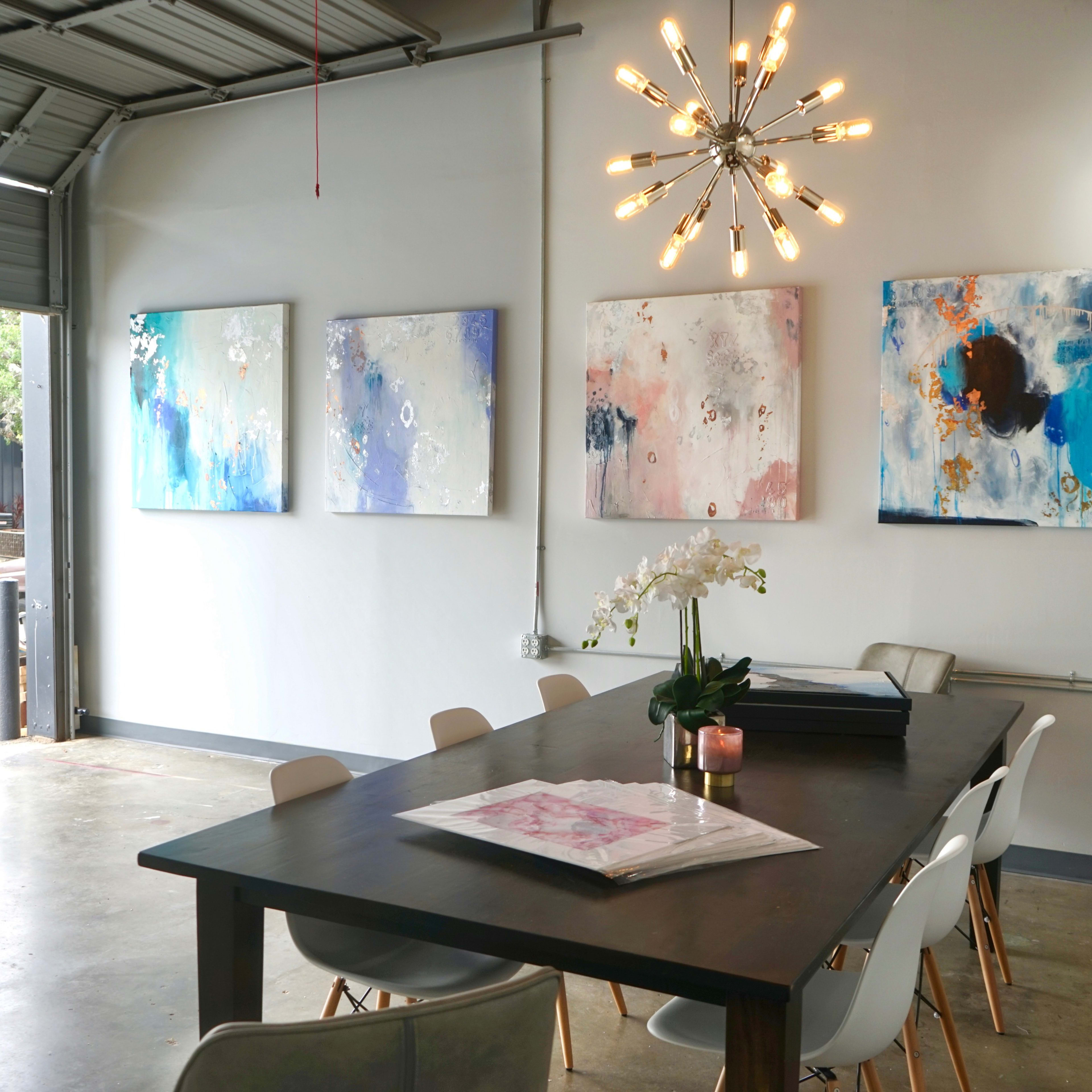 A modern meeting room with a large table, surrounded by chairs, and four colorful abstract paintings on the wall, illuminated by a unique light fixture.