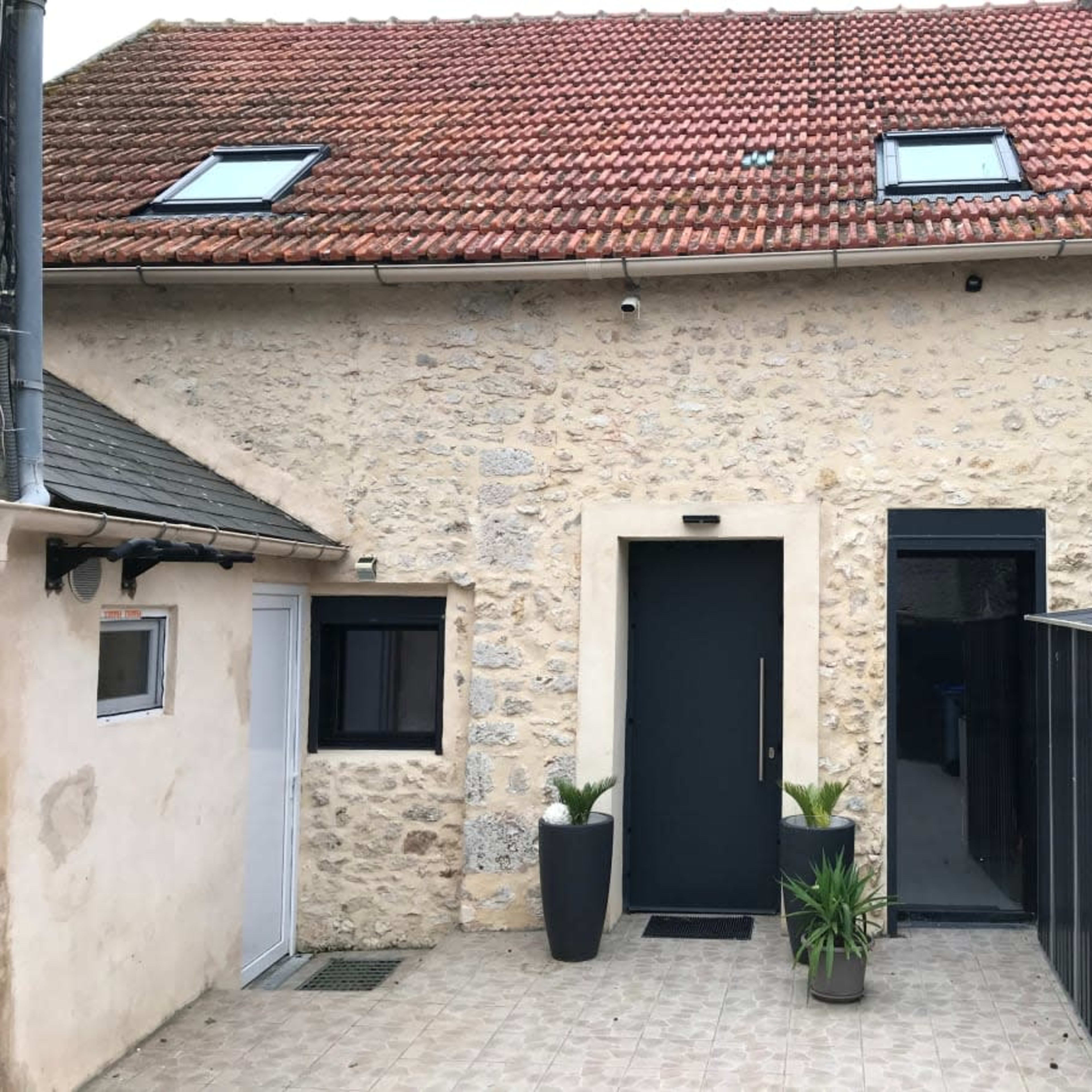 A stone building with a flat entrance door, flanked by two tall planters, and a dark siding storage area to the right.