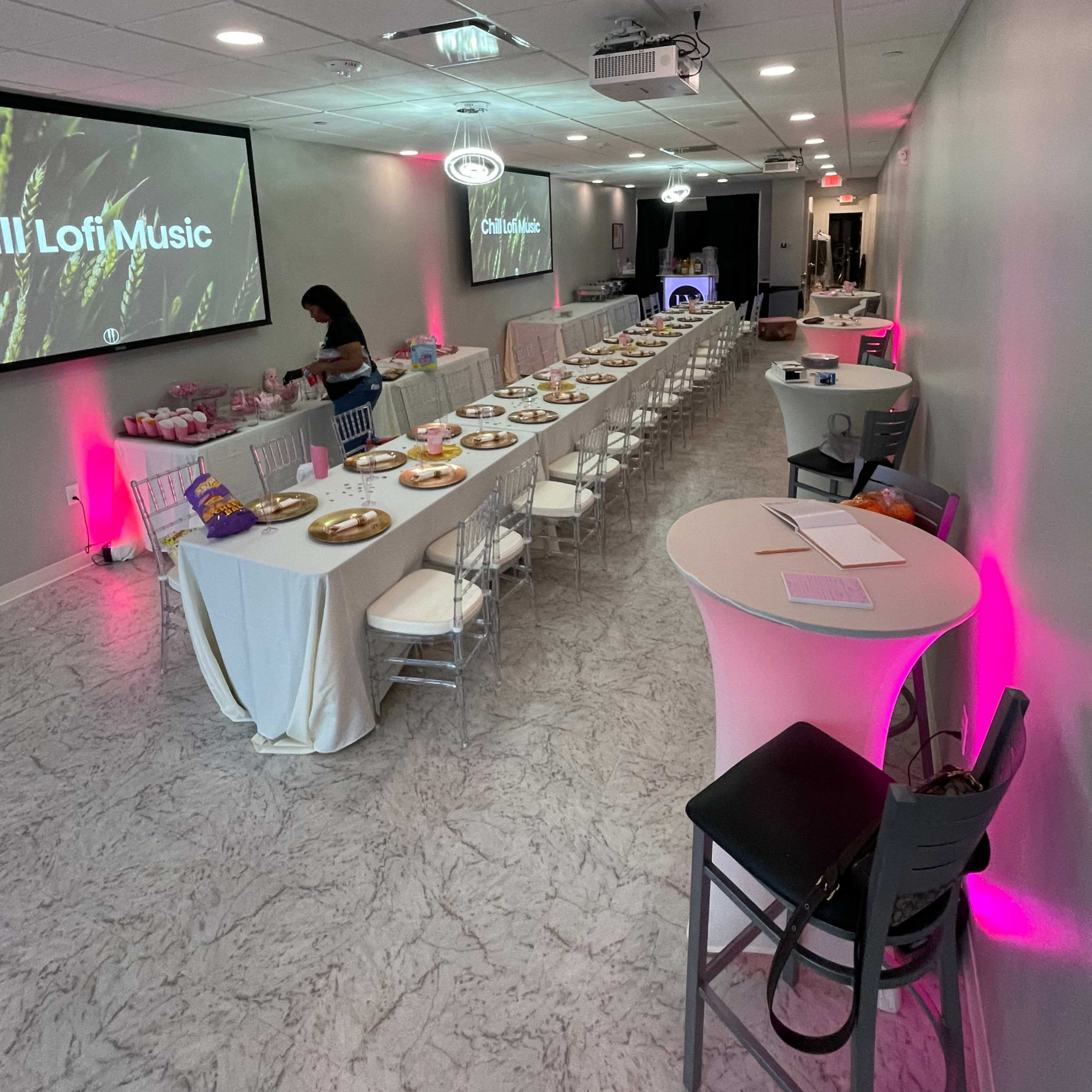 A modern event space set up with tables arranged in rows, featuring food platters, a large screen displaying "Chill Loft Music," and pink ambient lighting.