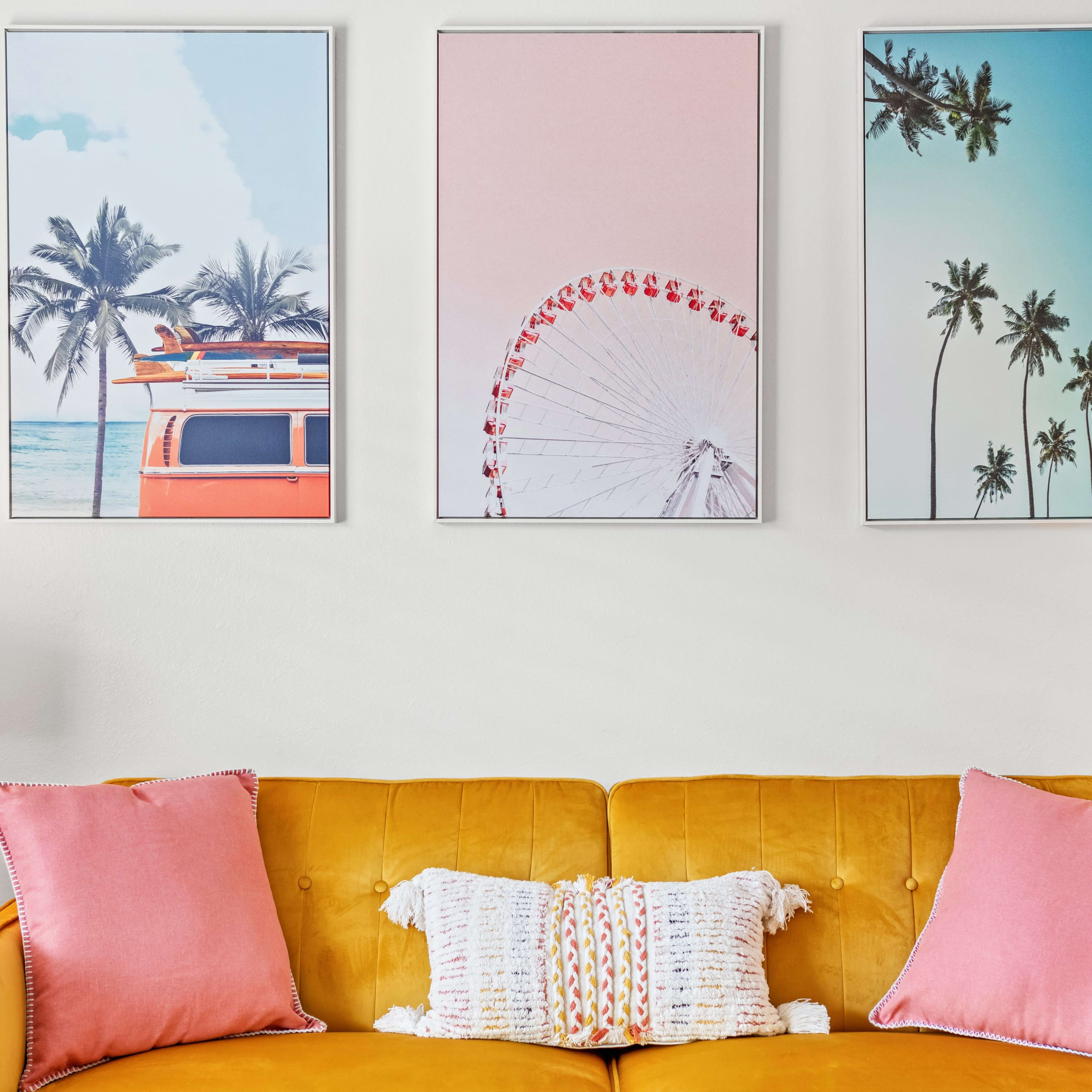 A yellow sofa with pink cushions is positioned below three framed landscape prints featuring a vintage van, a Ferris wheel, and palm trees.