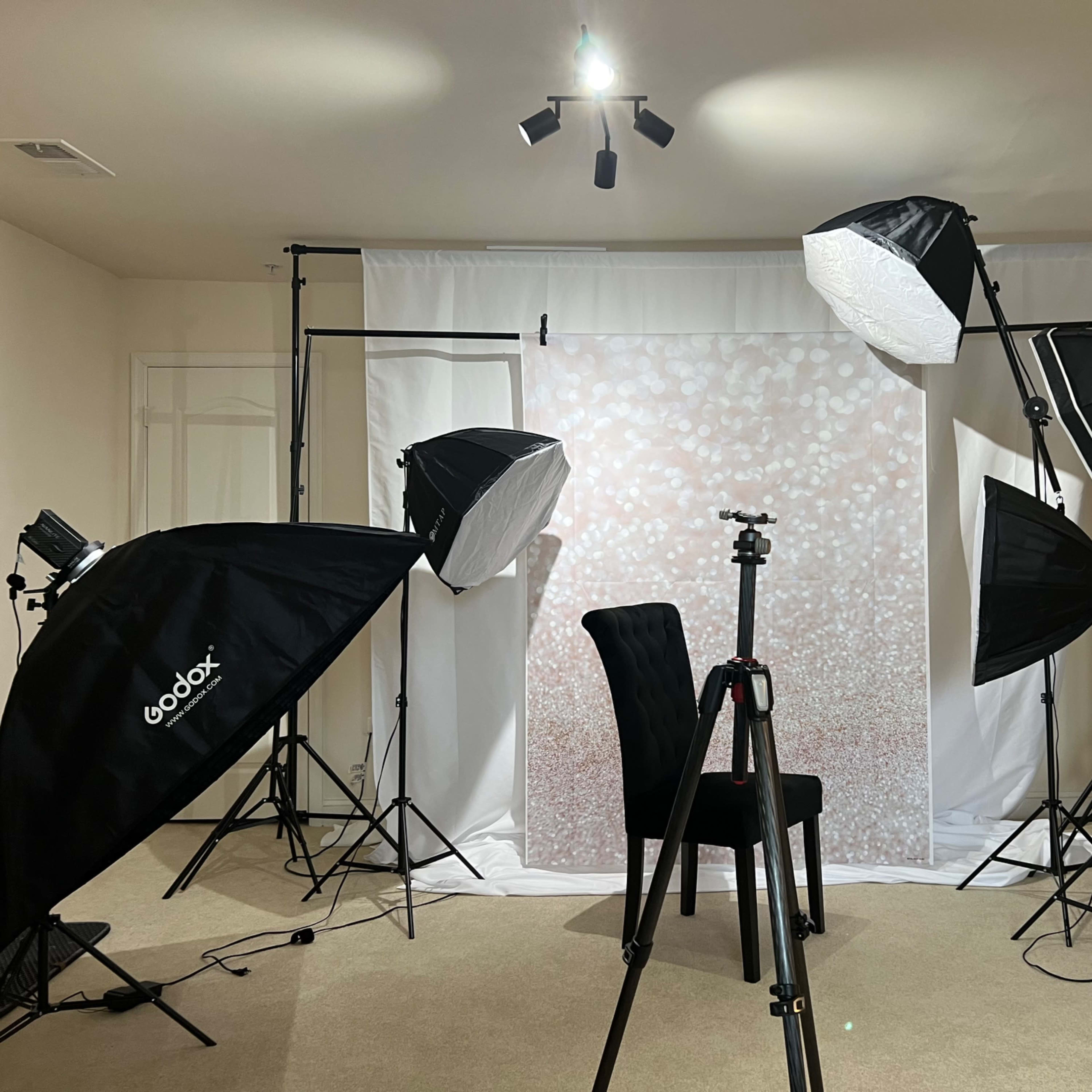 A photography studio setup featuring multiple softbox lights, a backdrop with a glittery design, a tripod, and a single chair positioned in the center.