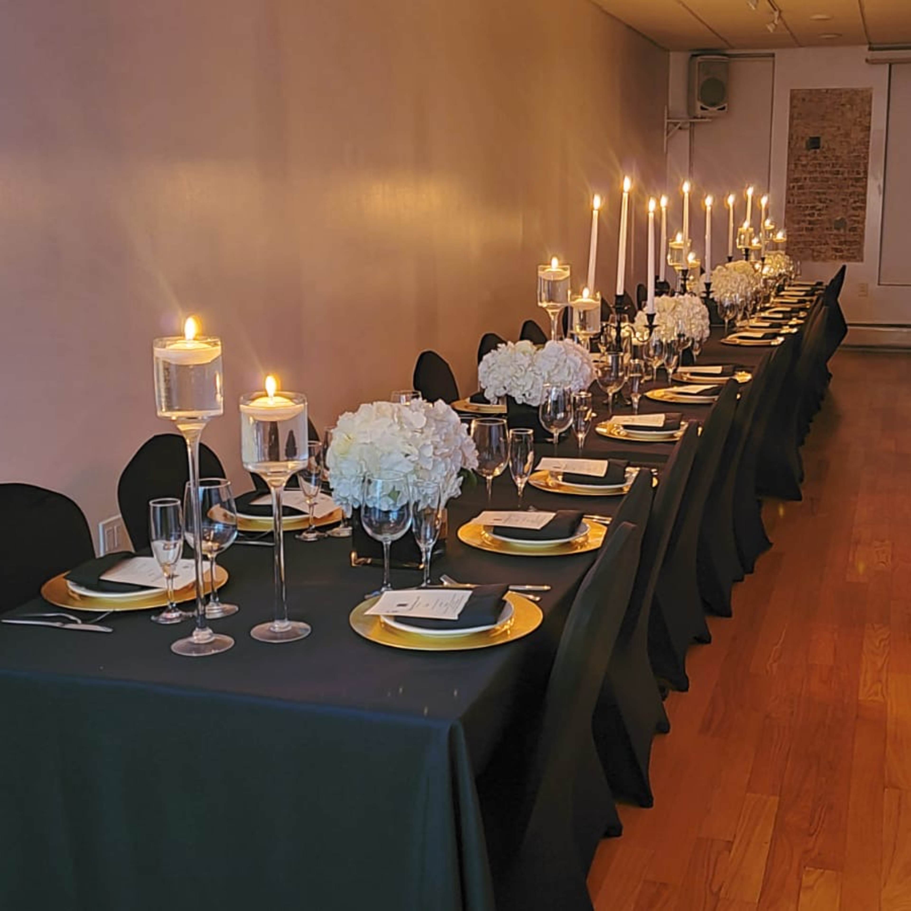 A long dining table set with gold plates, glassware, and lit candles, adorned with white floral centerpieces, is arranged in a spacious room with wooden flooring.