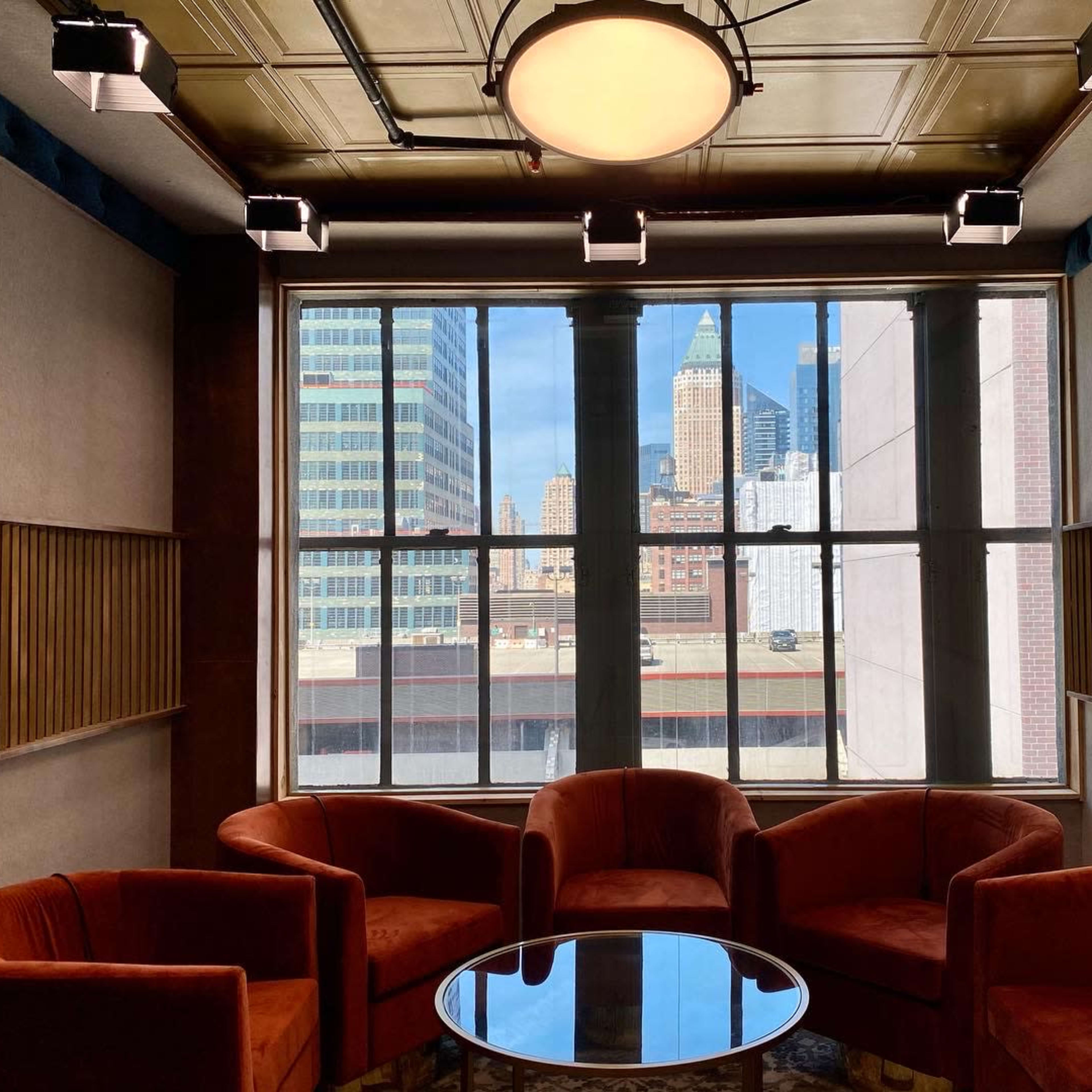 A seating area with four orange chairs arranged around a glass table, framed by large windows that overlook a cityscape featuring tall buildings.