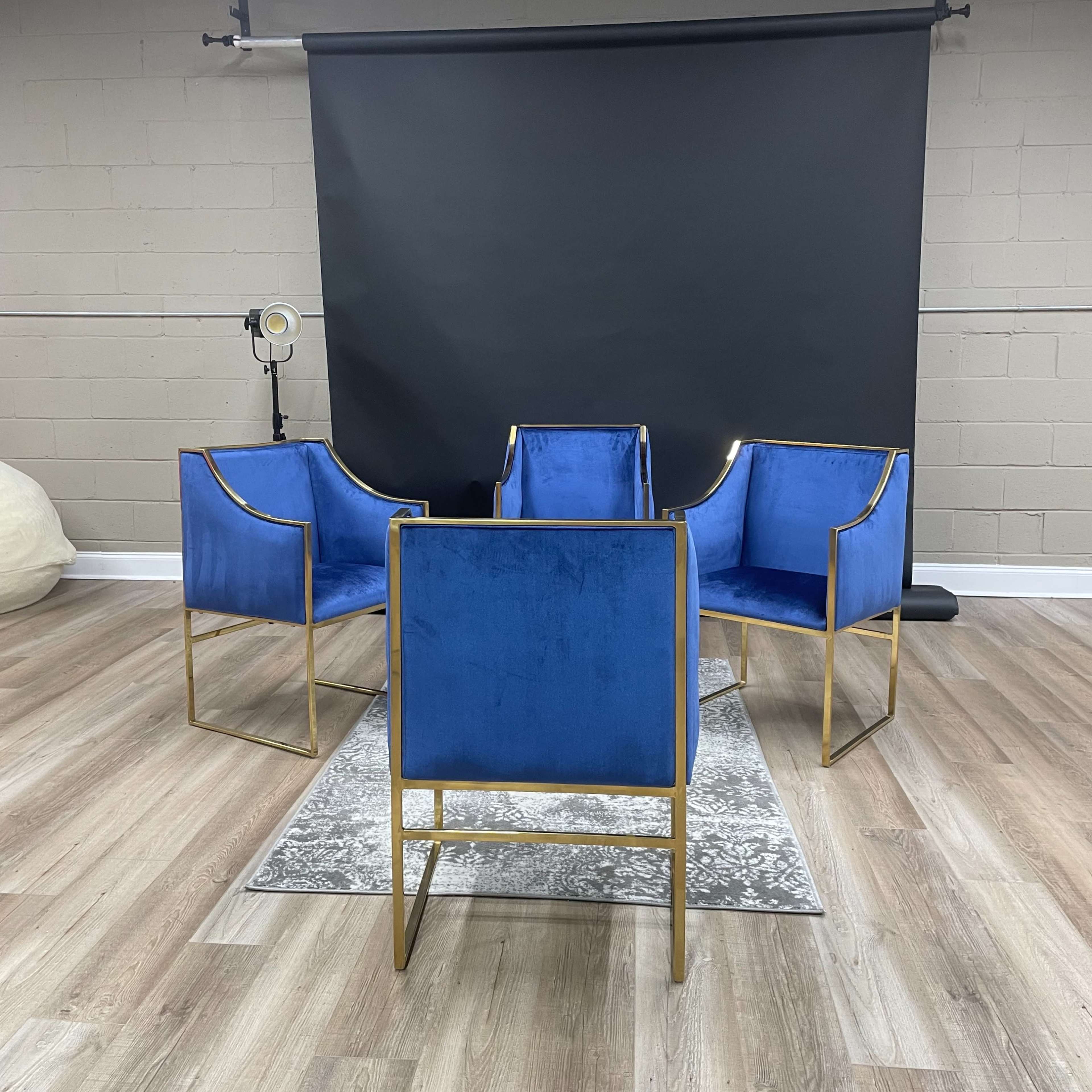 Four blue chairs with gold frames arranged around a patterned rug in a room with a black backdrop.
