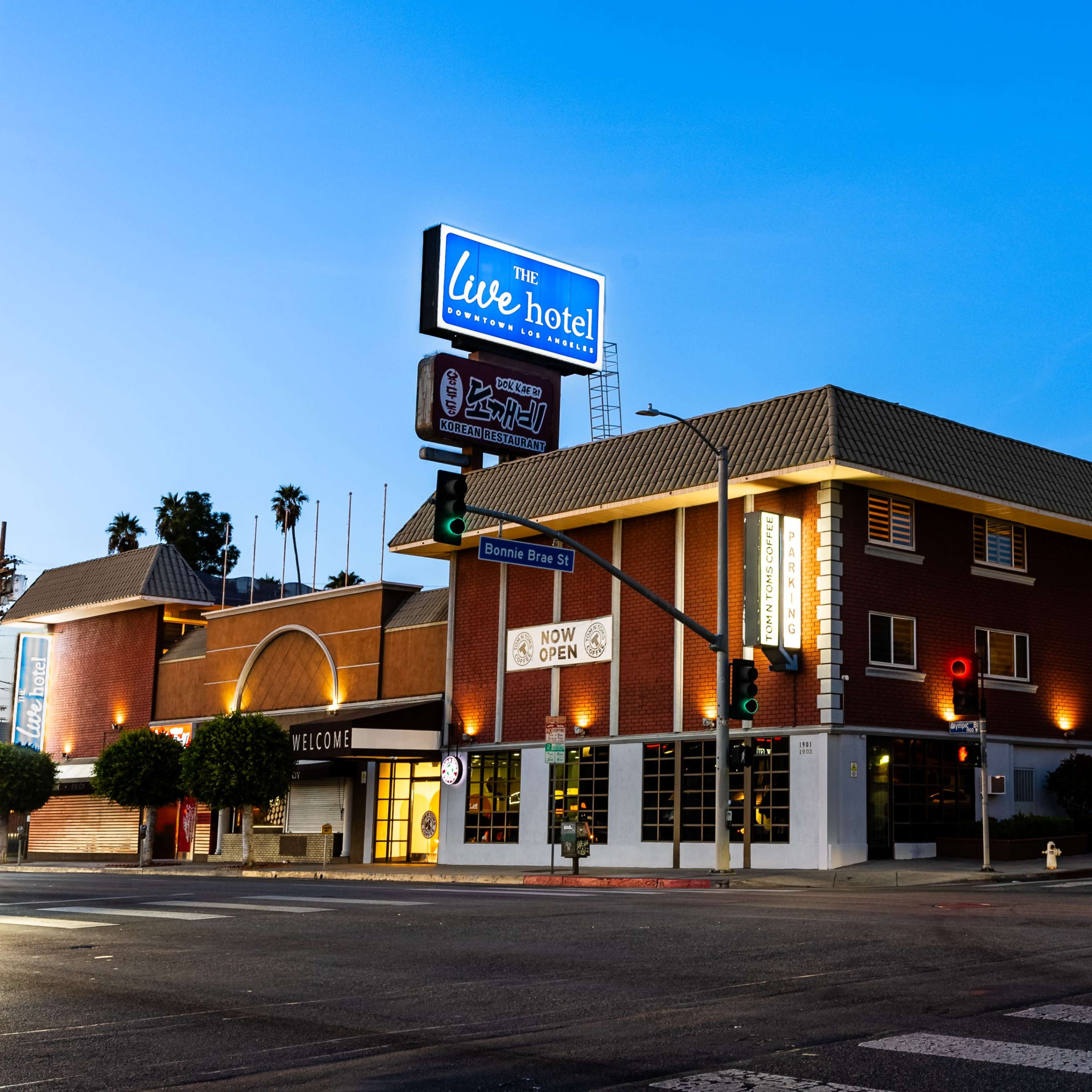 A brick hotel building at dusk, with illuminated signs and palm trees nearby, located at a street corner.