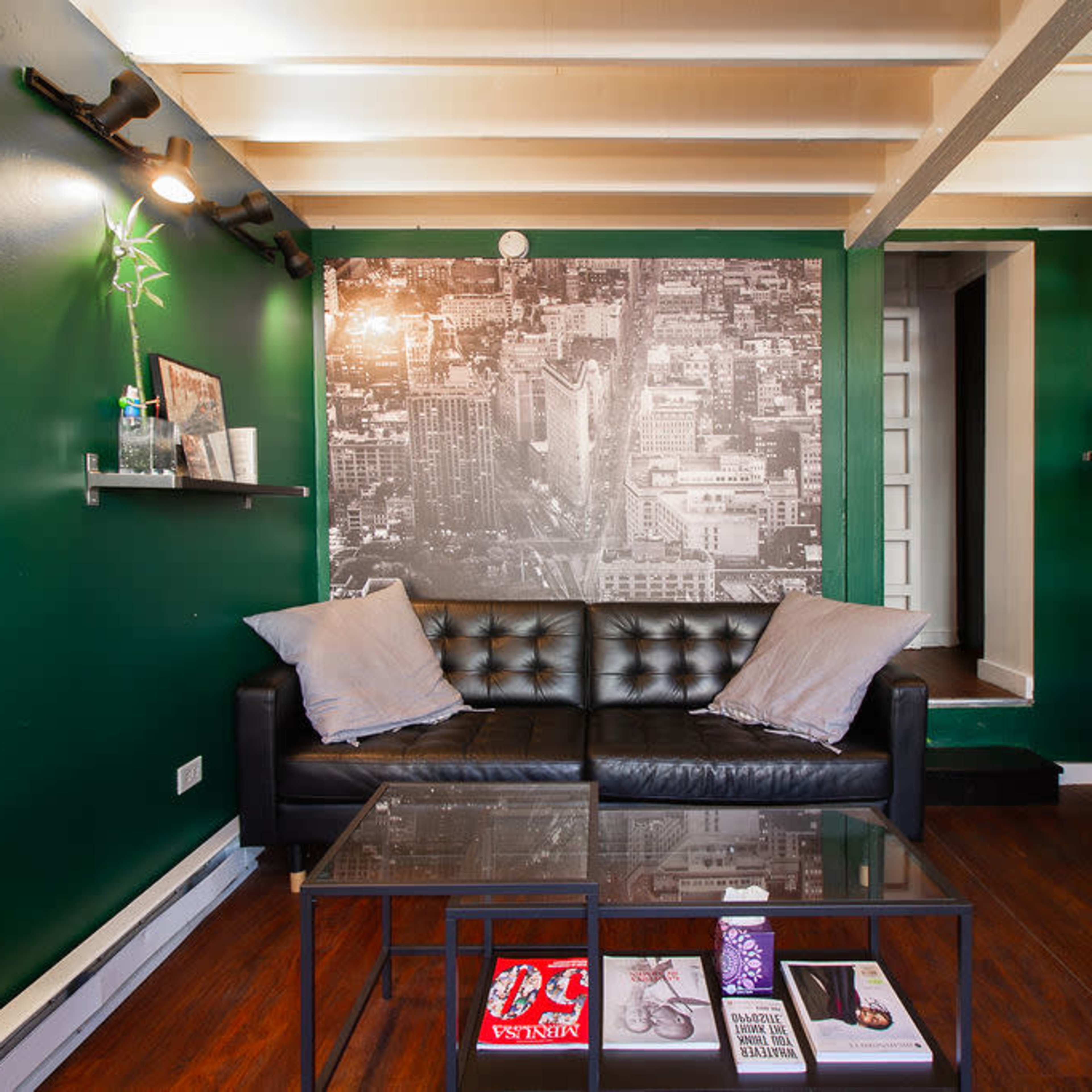 A small living space with dark green walls, a black leather couch, a coffee table with magazines, and shelves displaying various items.
