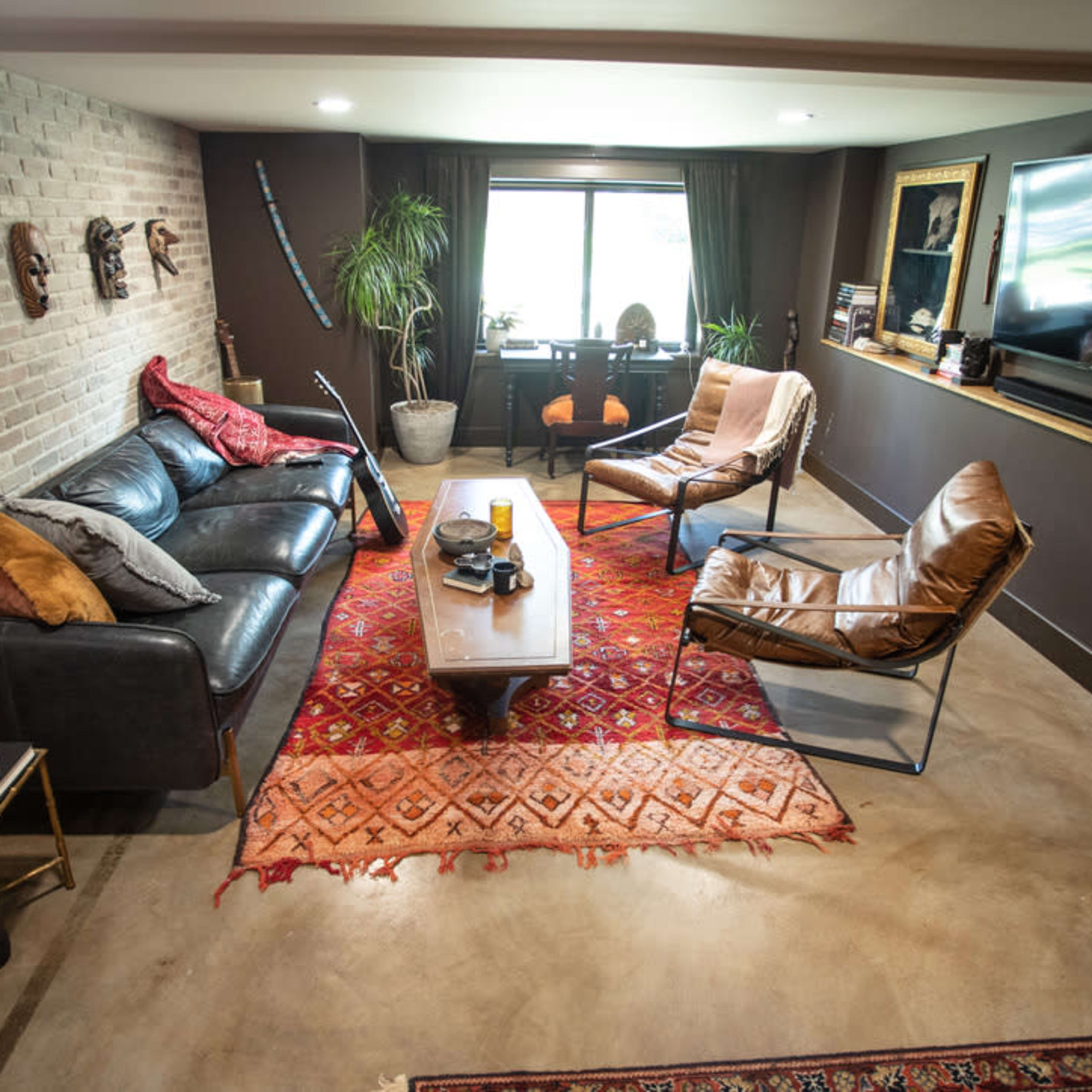 A modern living room featuring a black sofa, two leather chairs, a wooden coffee table on a patterned rug, and decorative masks on the brick wall.