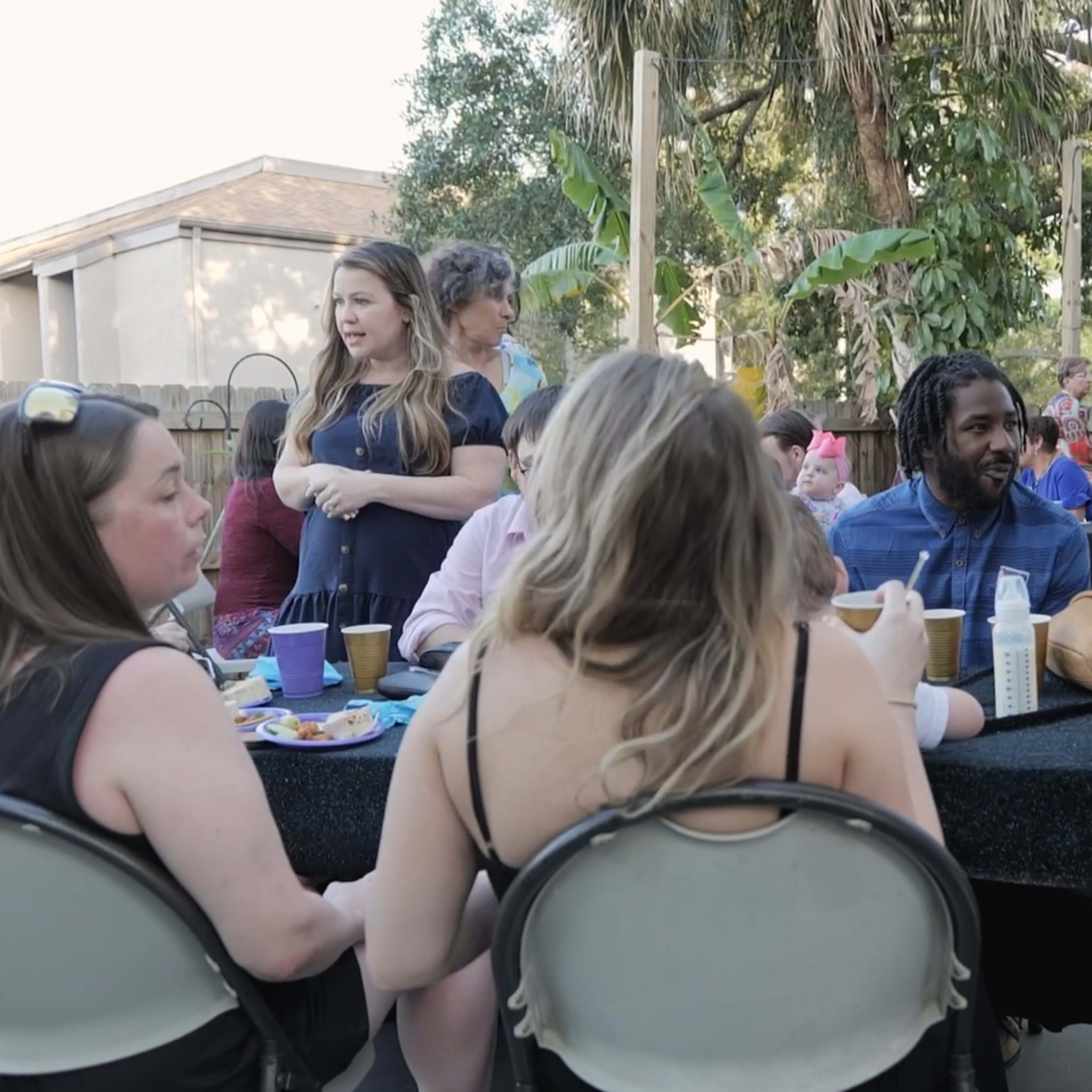 A gathering of people is taking place outdoors, with guests seated at tables and others mingling in a lively social atmosphere.