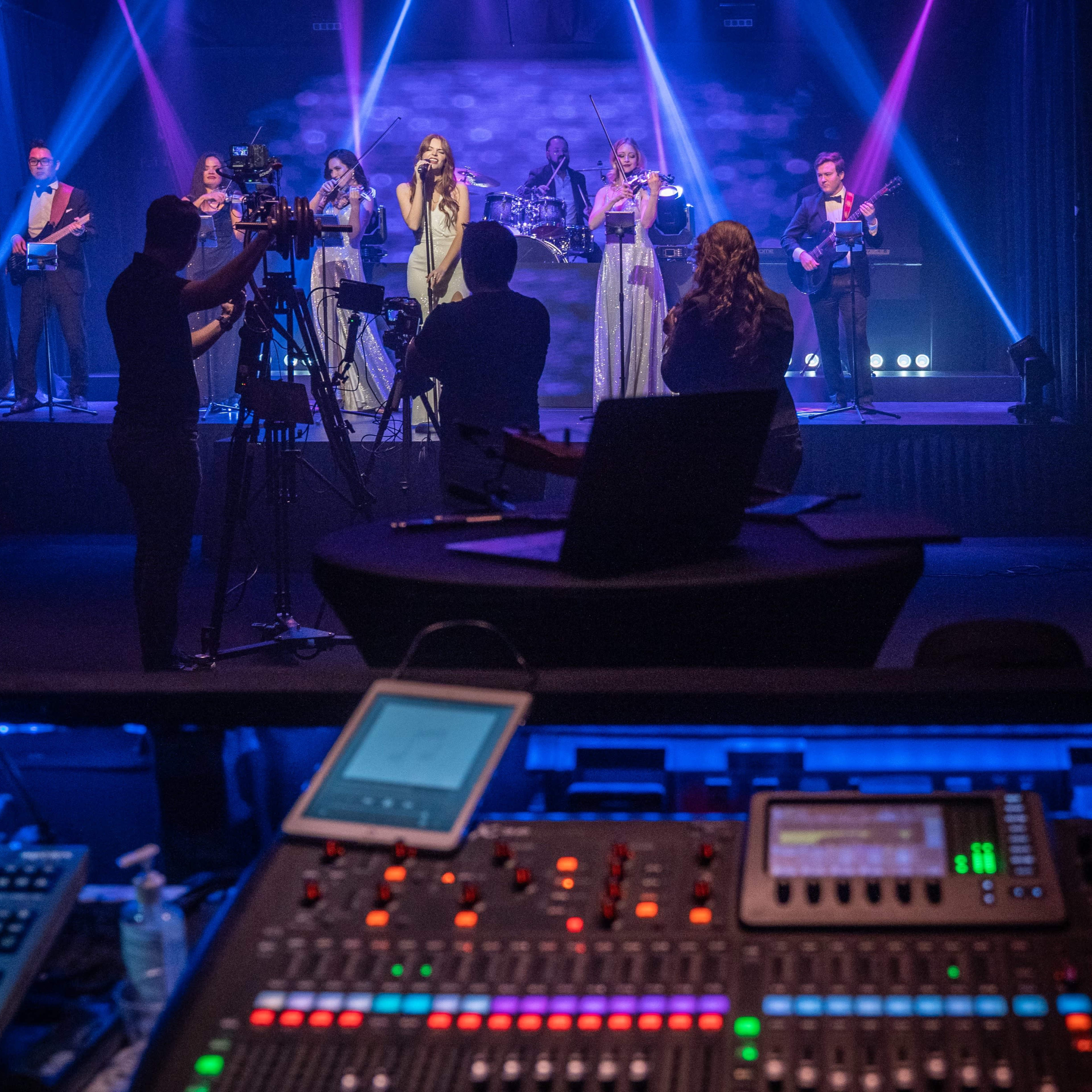 A live music performance is taking place on stage, with singers and musicians in formal attire and cameras capturing the event from the front.