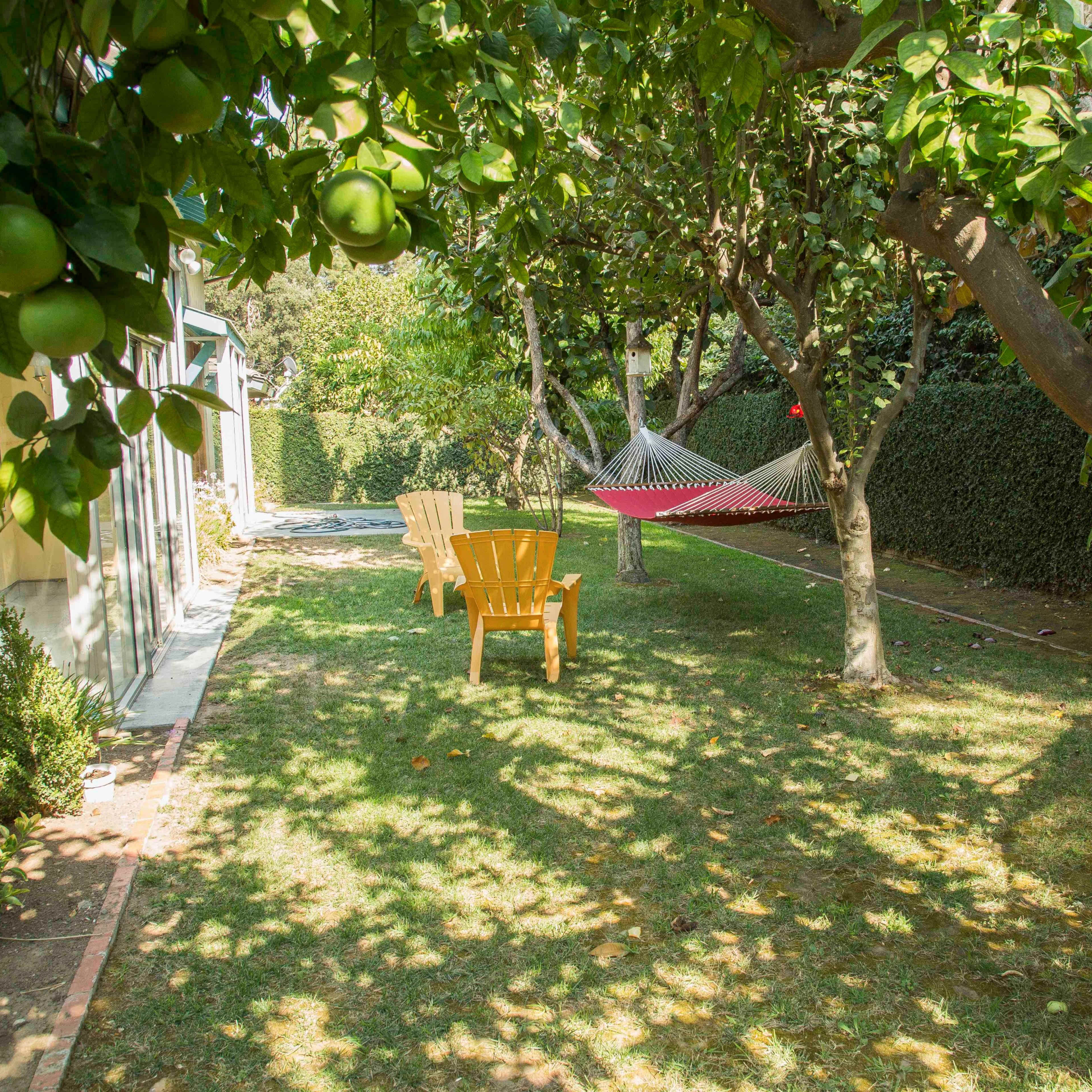 A backyard features a green lawn with a hammock strung between trees and two yellow chairs nearby.