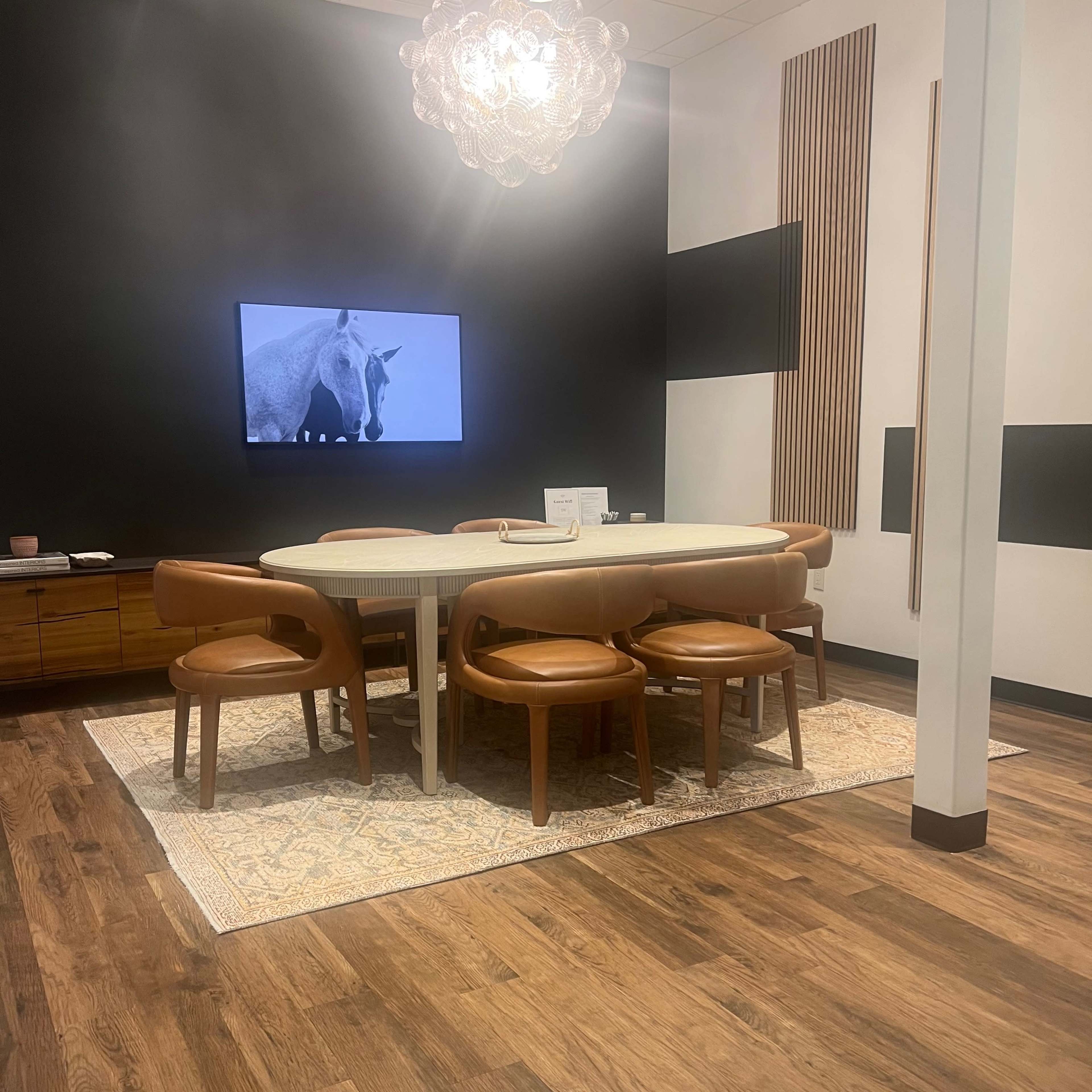 A modern conference room features a rectangular table surrounded by leather chairs, a large screen displaying a horse image, and a pendant light hanging from the ceiling.