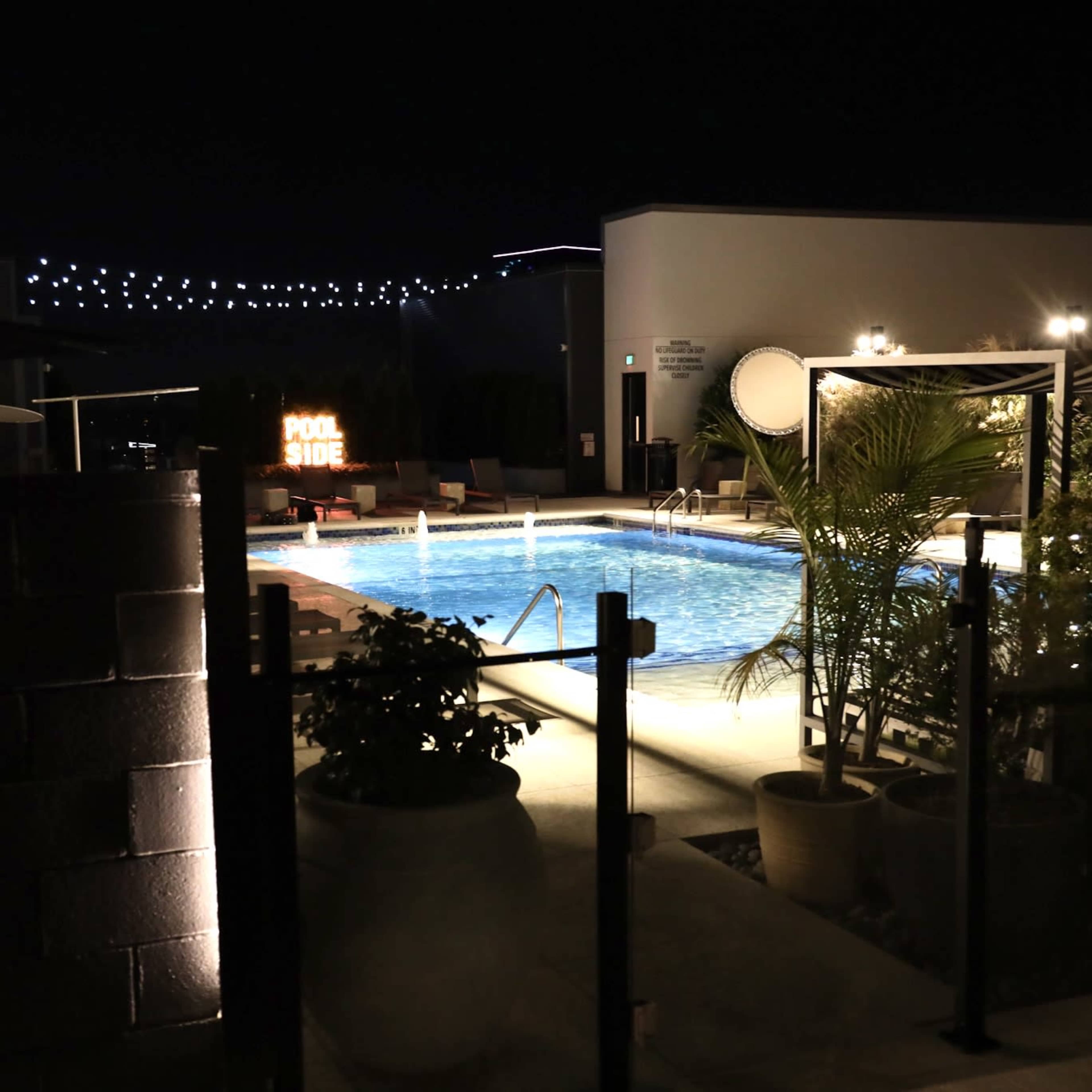 A well-lit swimming pool surrounded by lounge chairs and greenery is visible at night.