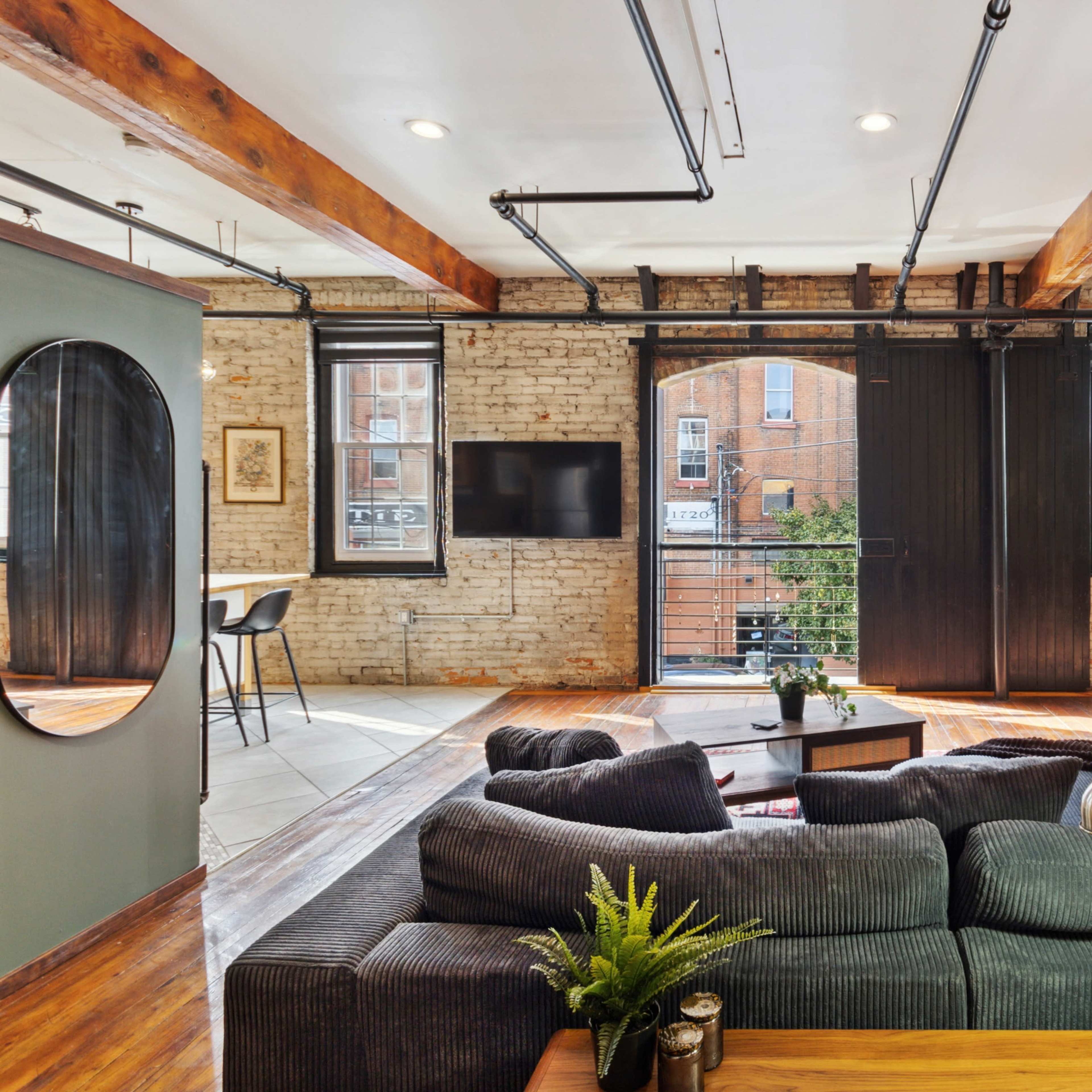 A spacious living area featuring exposed brick walls, wooden beams, large windows, and a mix of modern and vintage decor.