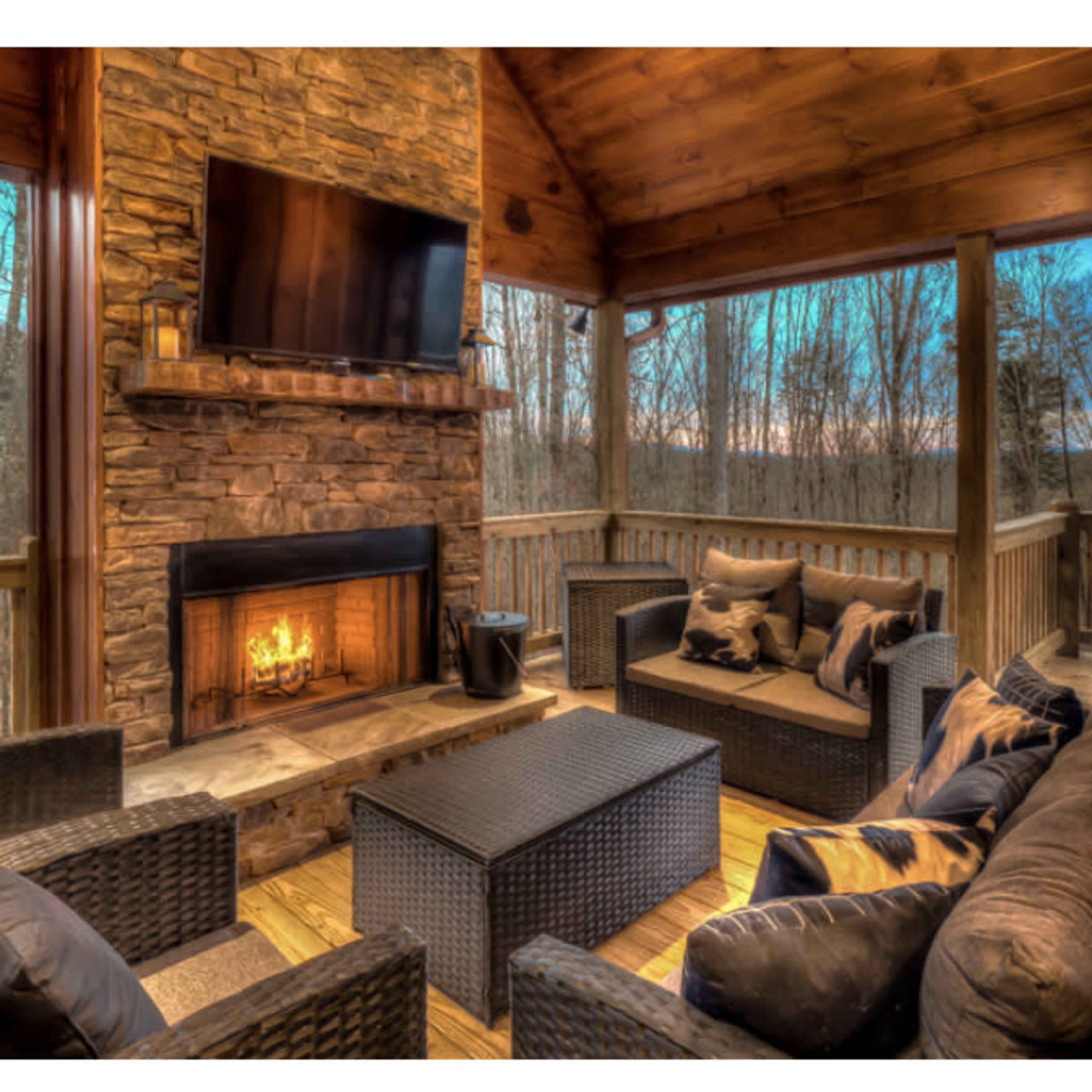 A wooden porch with a stone fireplace, seating area, and a television, surrounded by trees at dusk.
