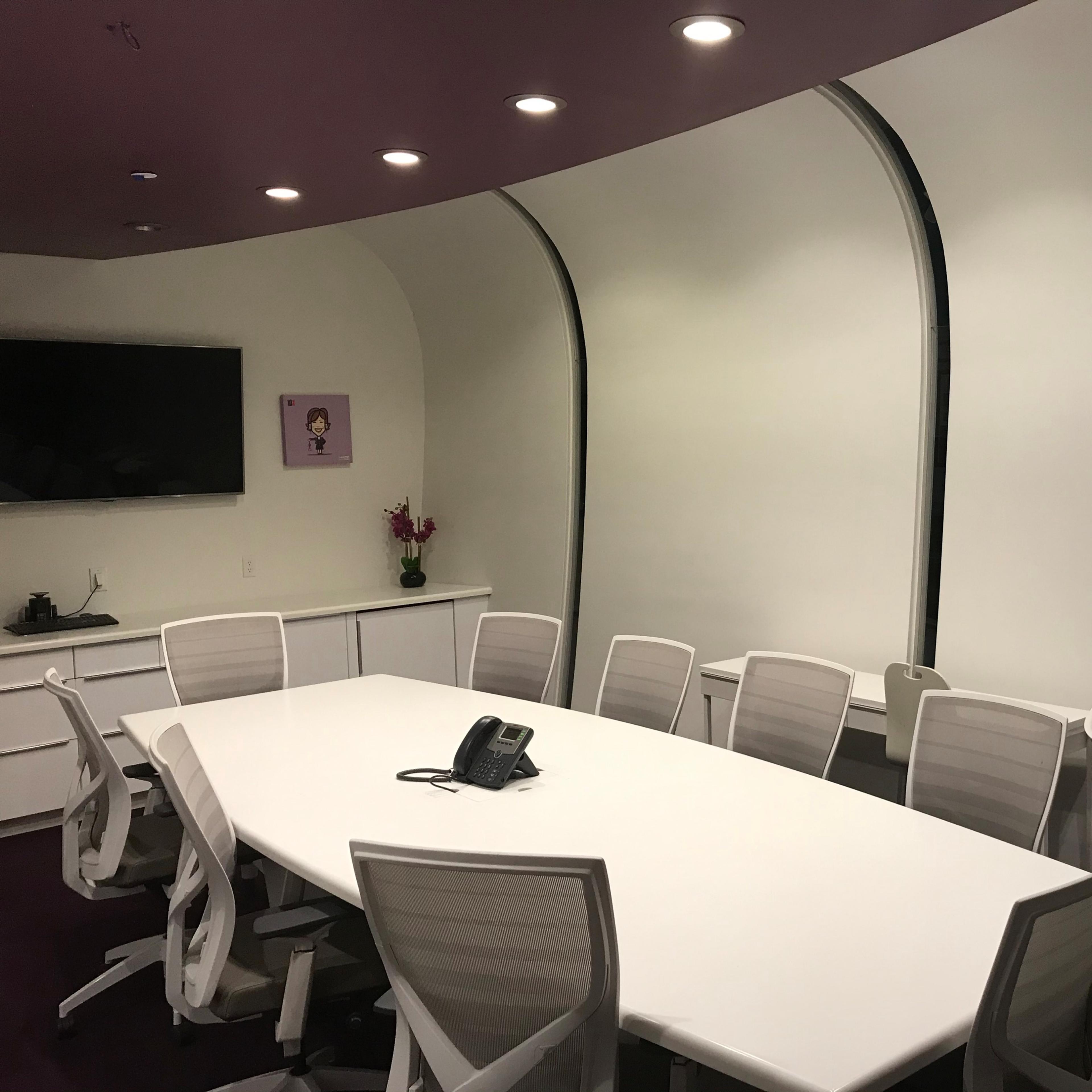 A modern conference room features a long white table, ergonomic chairs, a television, and a curved wall.
