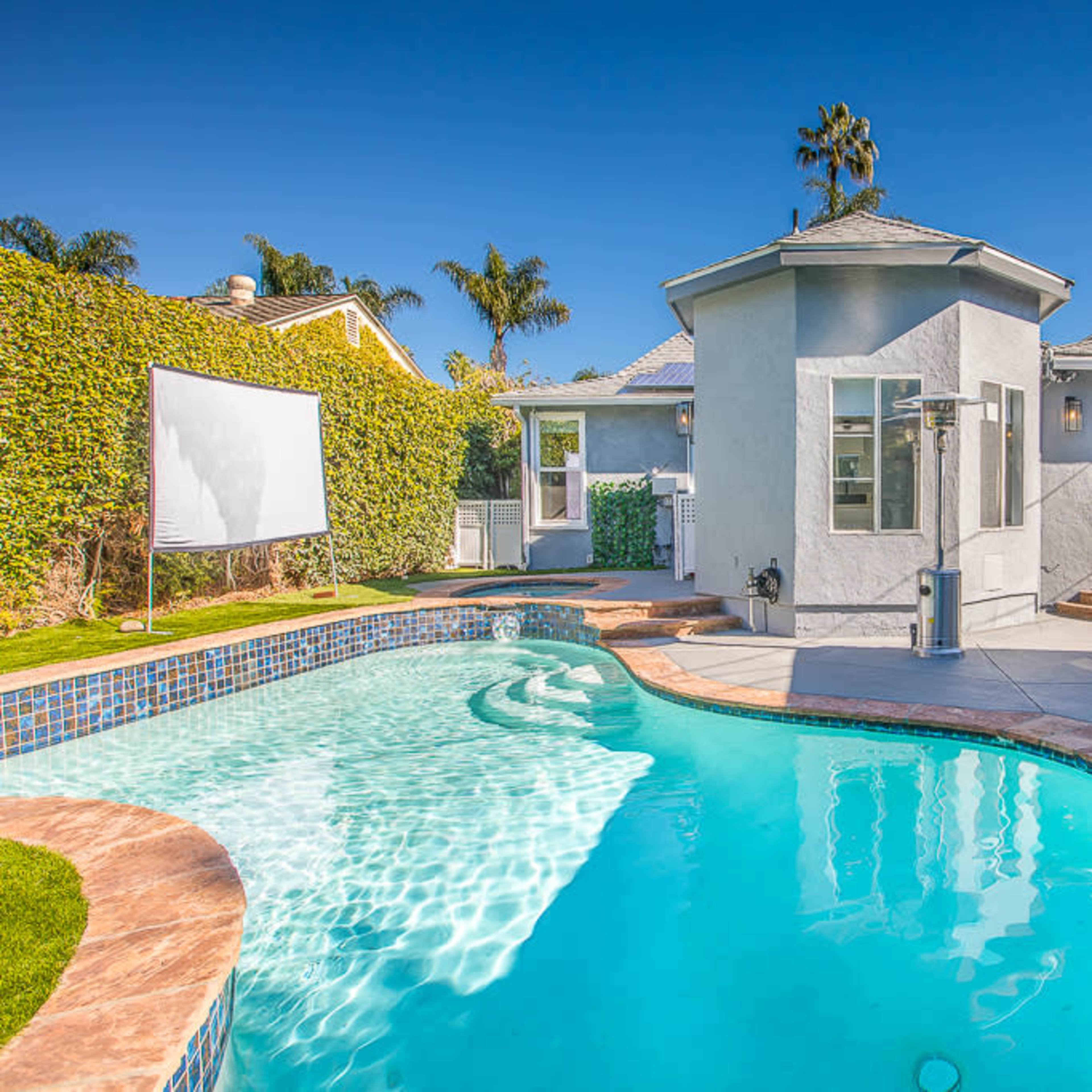 A residential backyard features a swimming pool with a stone edge, a cinema screen, and a small house with a porch.