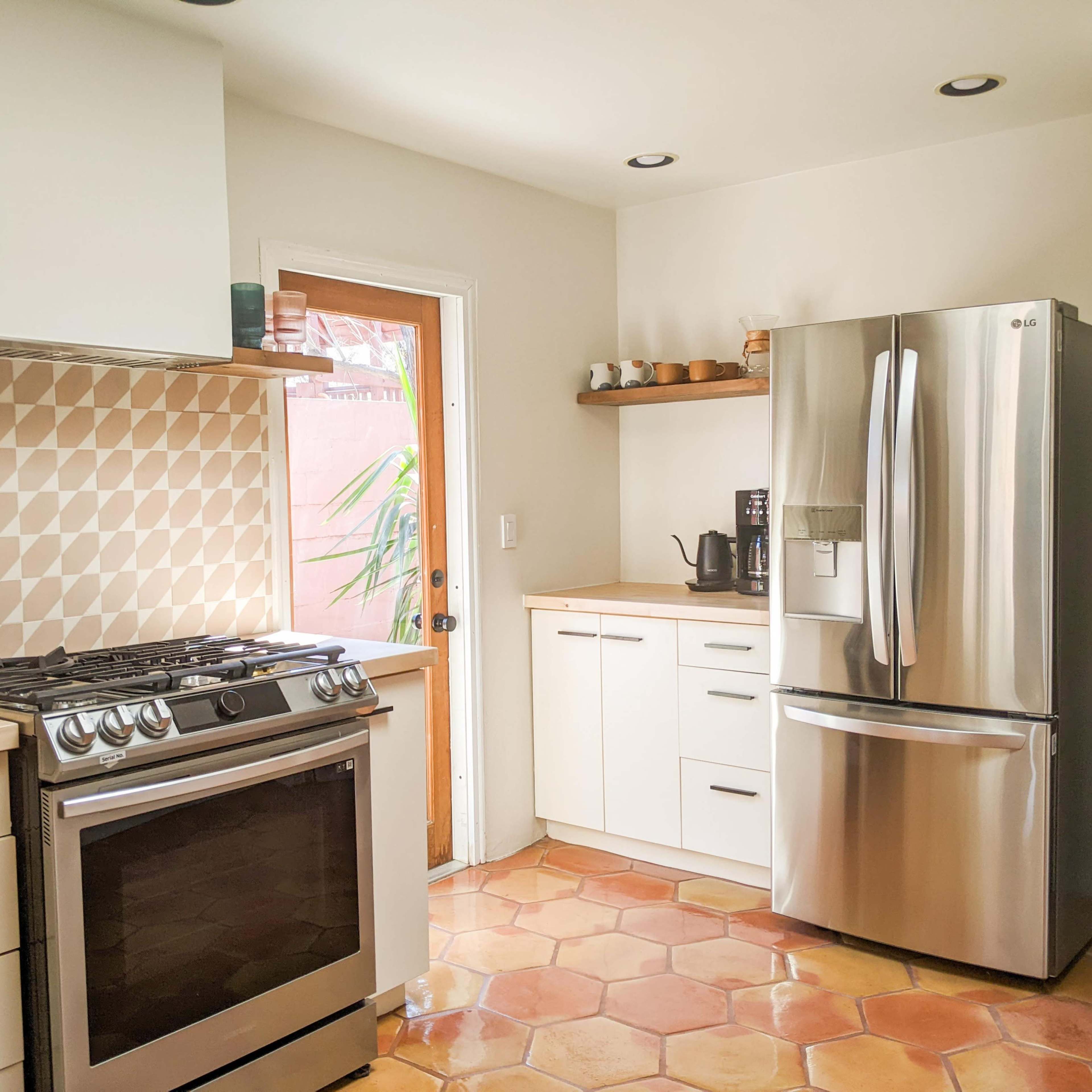 A modern kitchen with a stainless steel refrigerator, a gas stove, and light-colored cabinetry, featuring a textured backsplash and terra-cotta floor tiles.