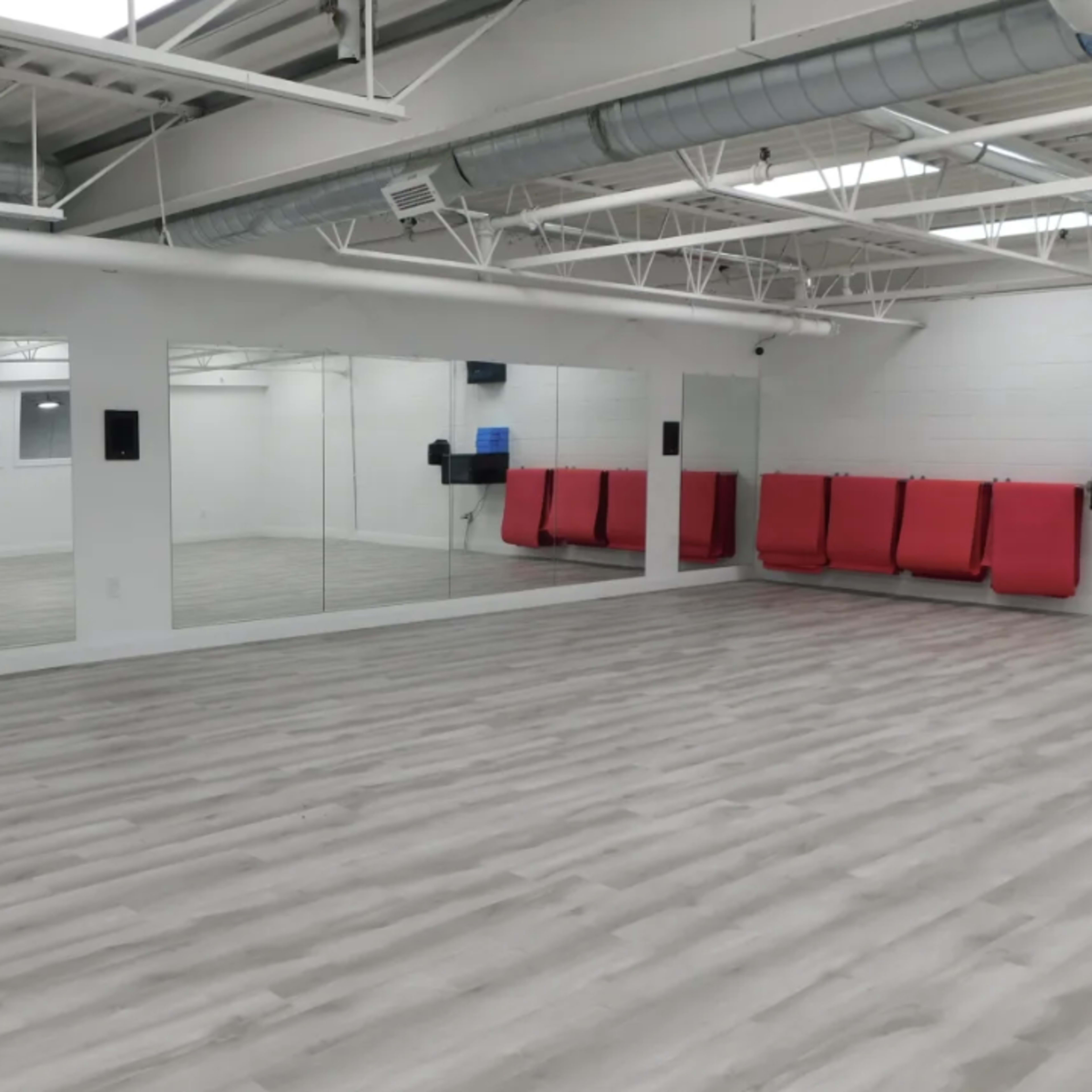 A spacious, empty studio with light-colored wooden flooring, large mirrors on one wall, and red seating against another wall.