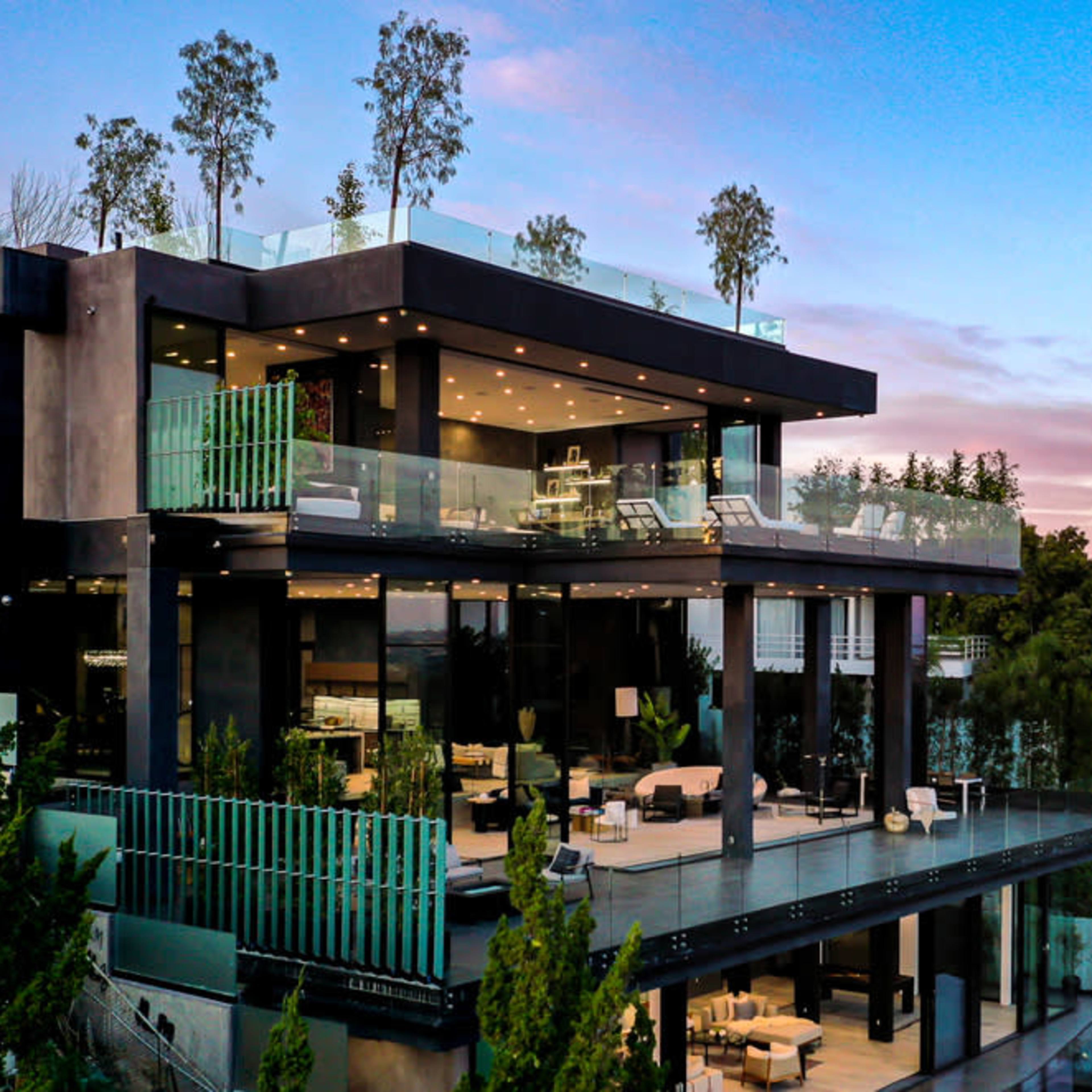 A modern multi-level house with glass balconies is situated on a hillside, surrounded by trees and overlooking distant mountains at sunset.