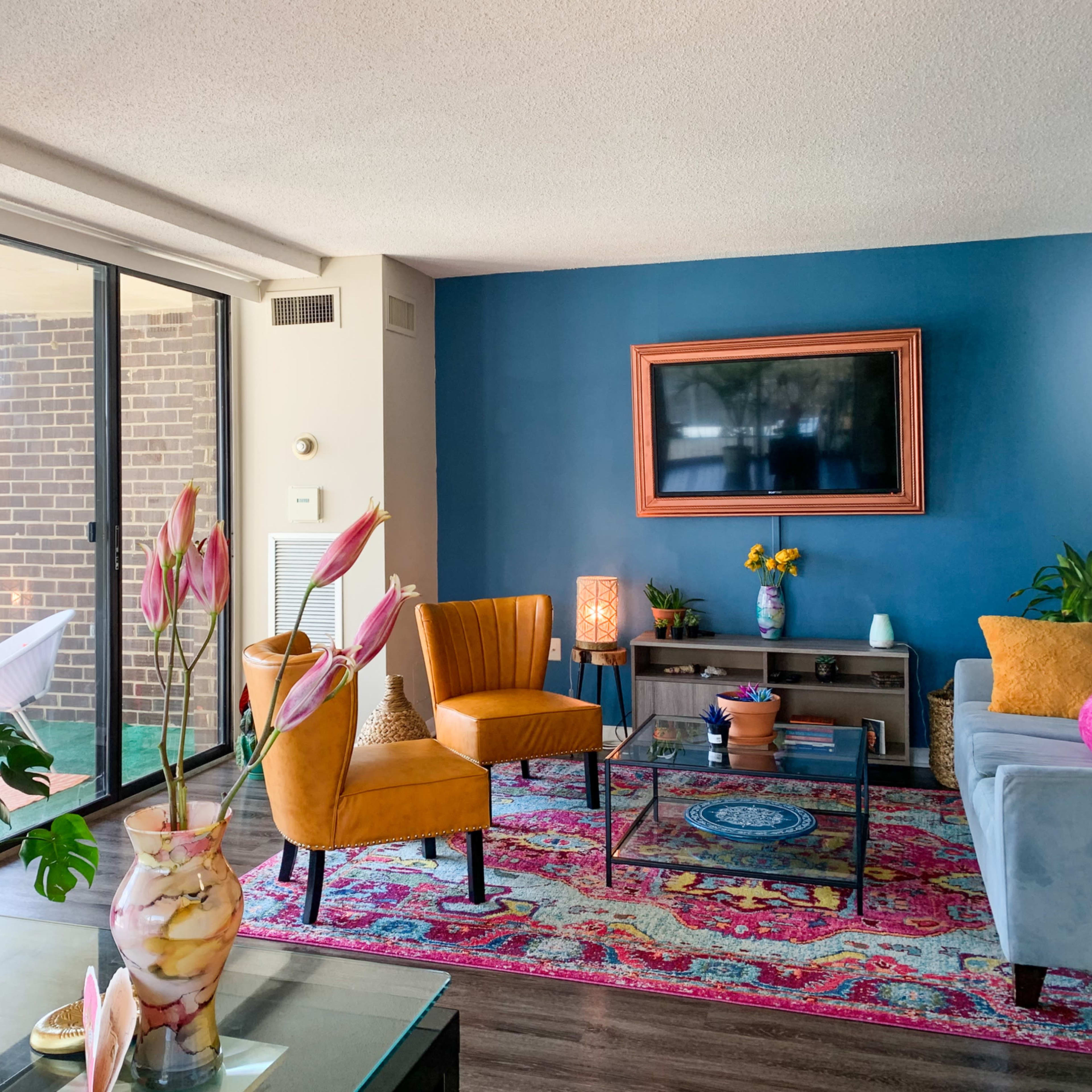 A colorful living room features a blue accent wall, a flat-screen TV, orange chairs, and a patterned rug.