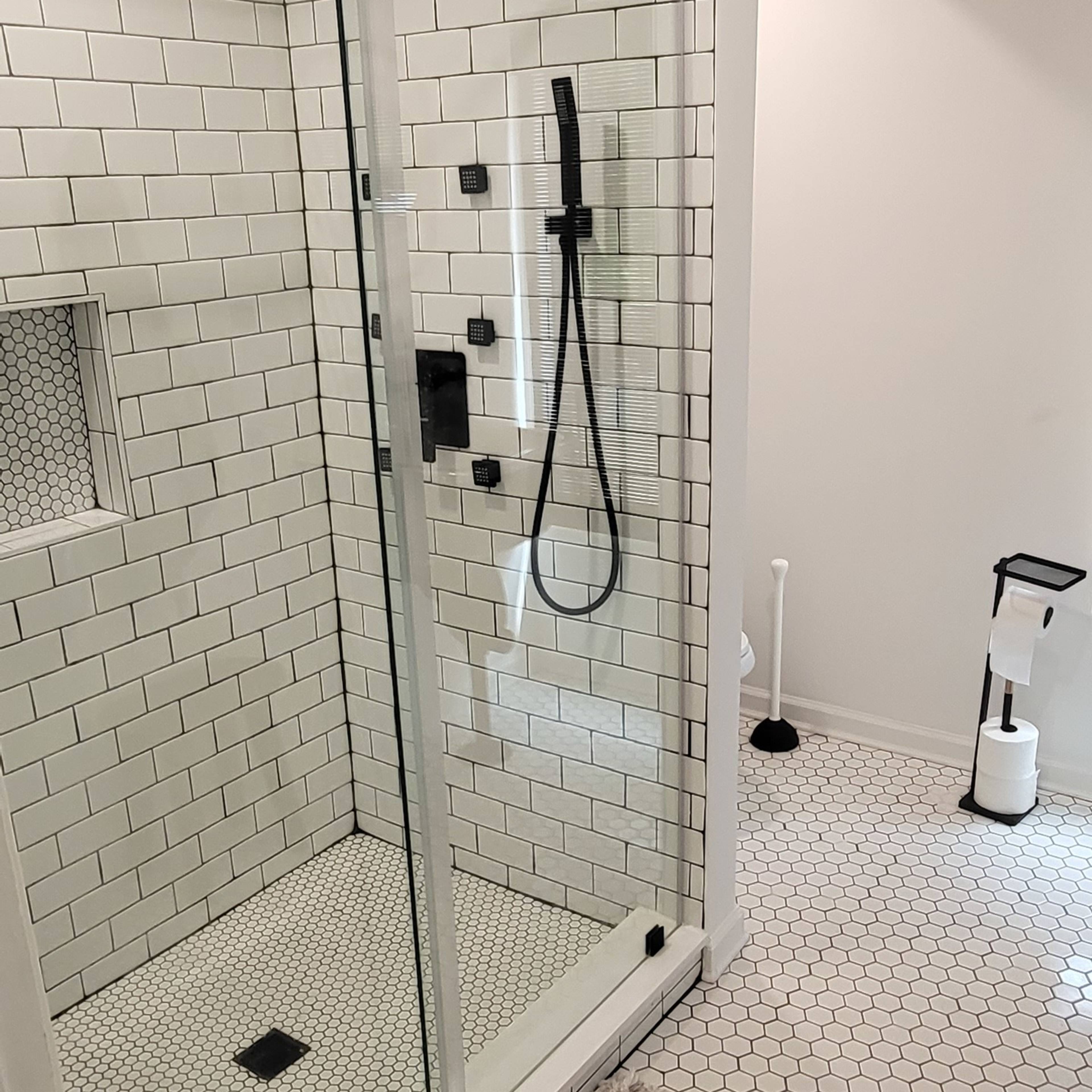 A modern bathroom featuring a glass shower enclosure with black fixtures, a hexagon tiled floor, and a light-colored rug.
