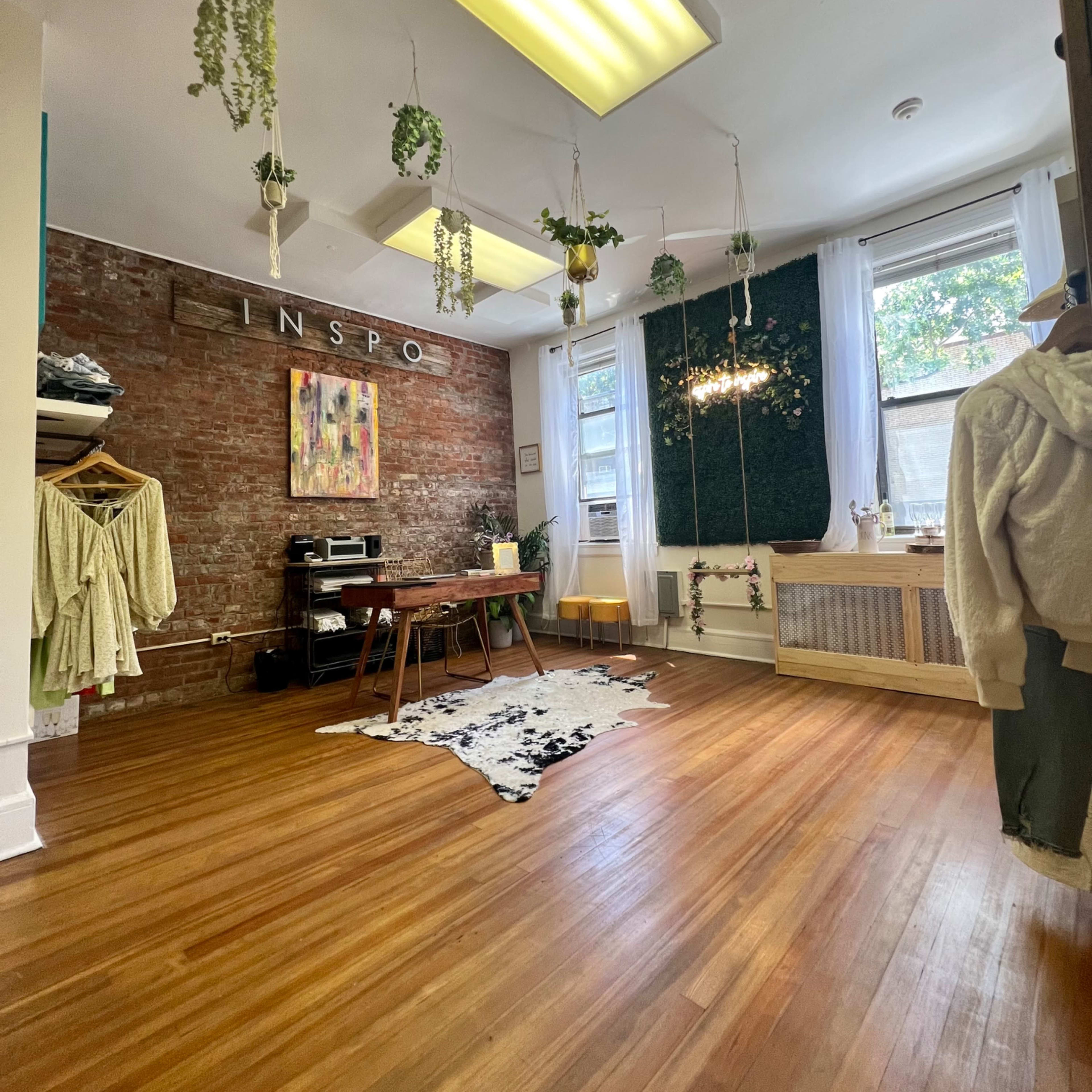 A spacious room with hardwood flooring, featuring a brick wall, hanging plants, and a mix of furniture including a table and clothing racks.