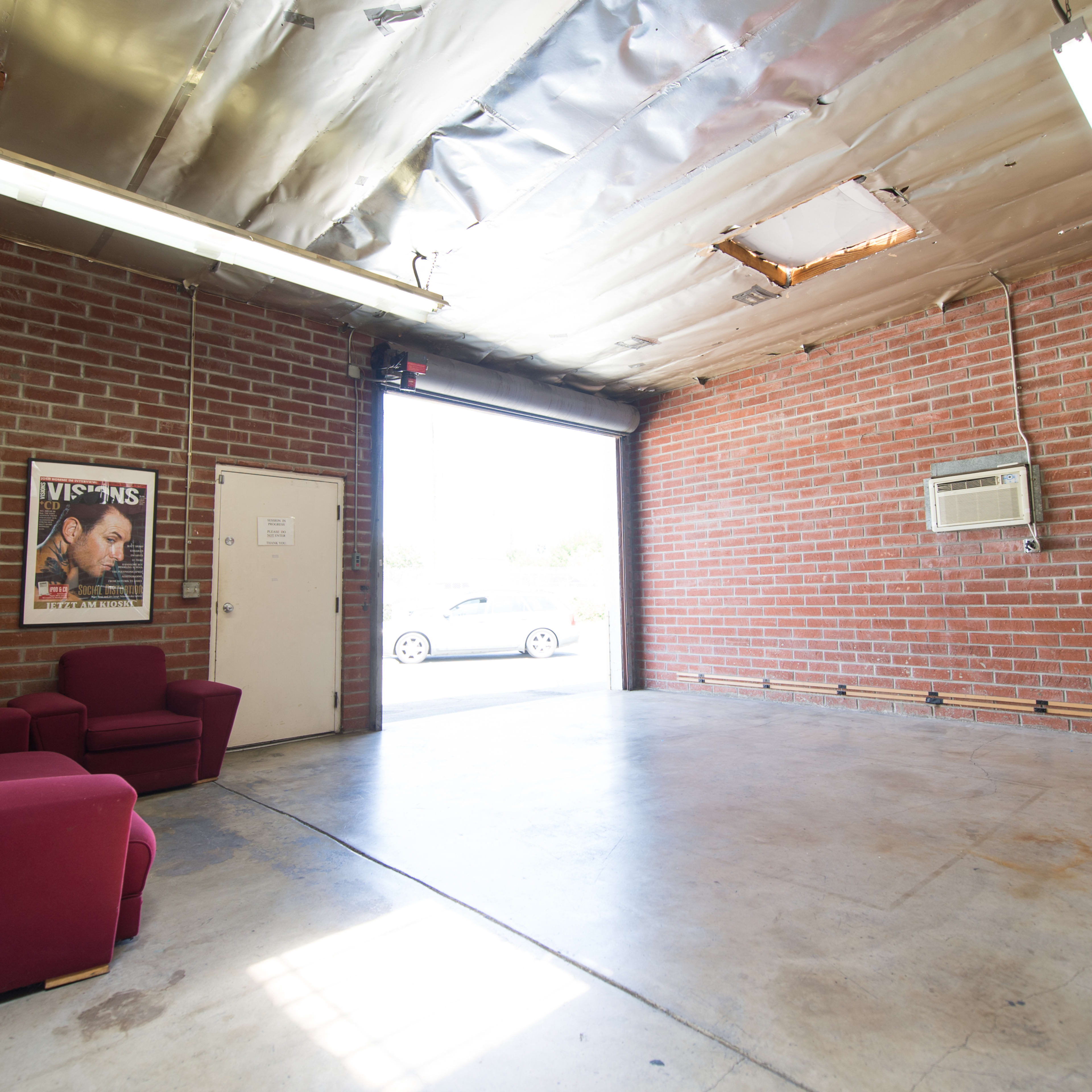 An empty garage with exposed brick walls, a ceiling with metal tiles, two red chairs, and a door leading outside.