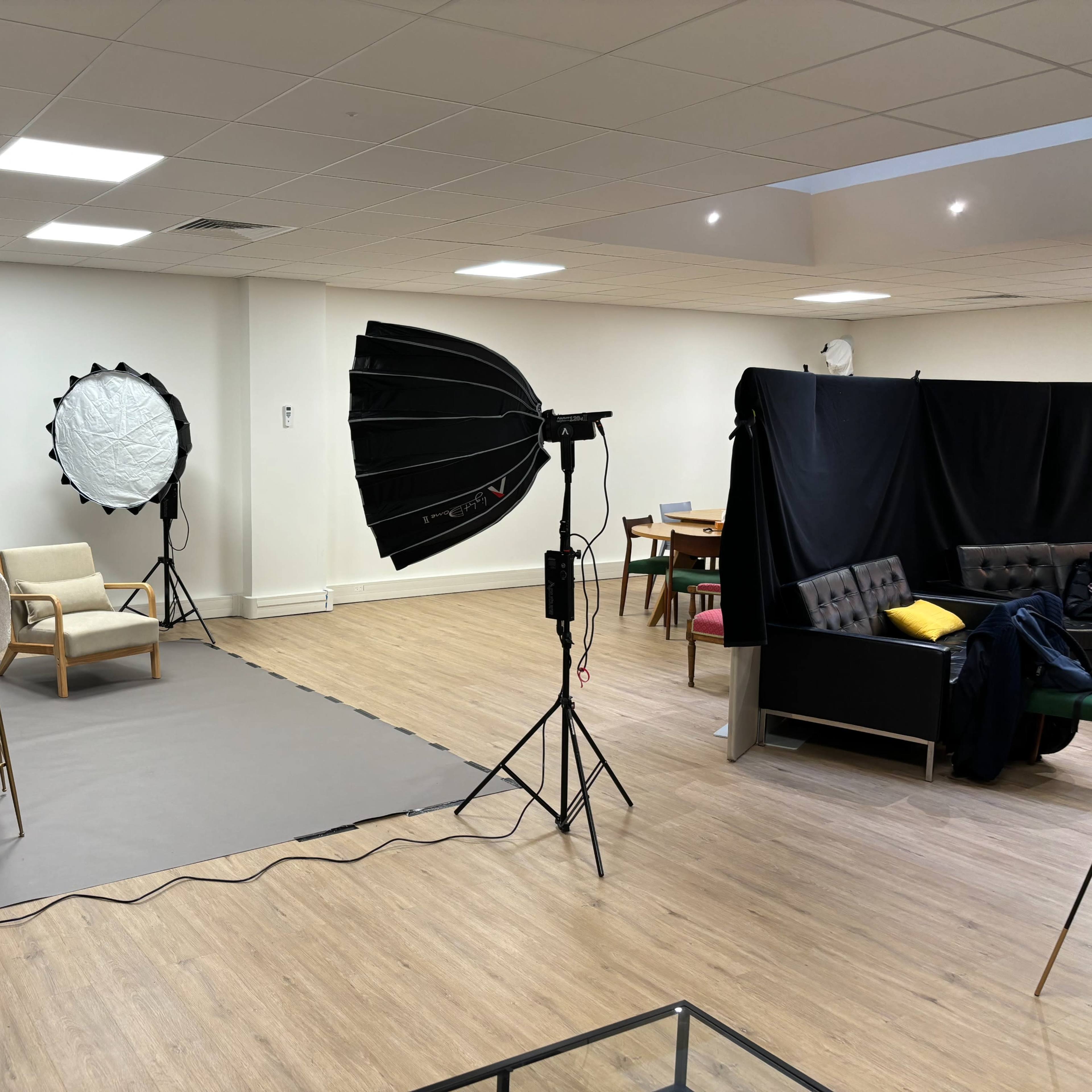 A well-lit studio setup featuring a seating area with chairs, lighting equipment, and a backdrop screen in a spacious room.