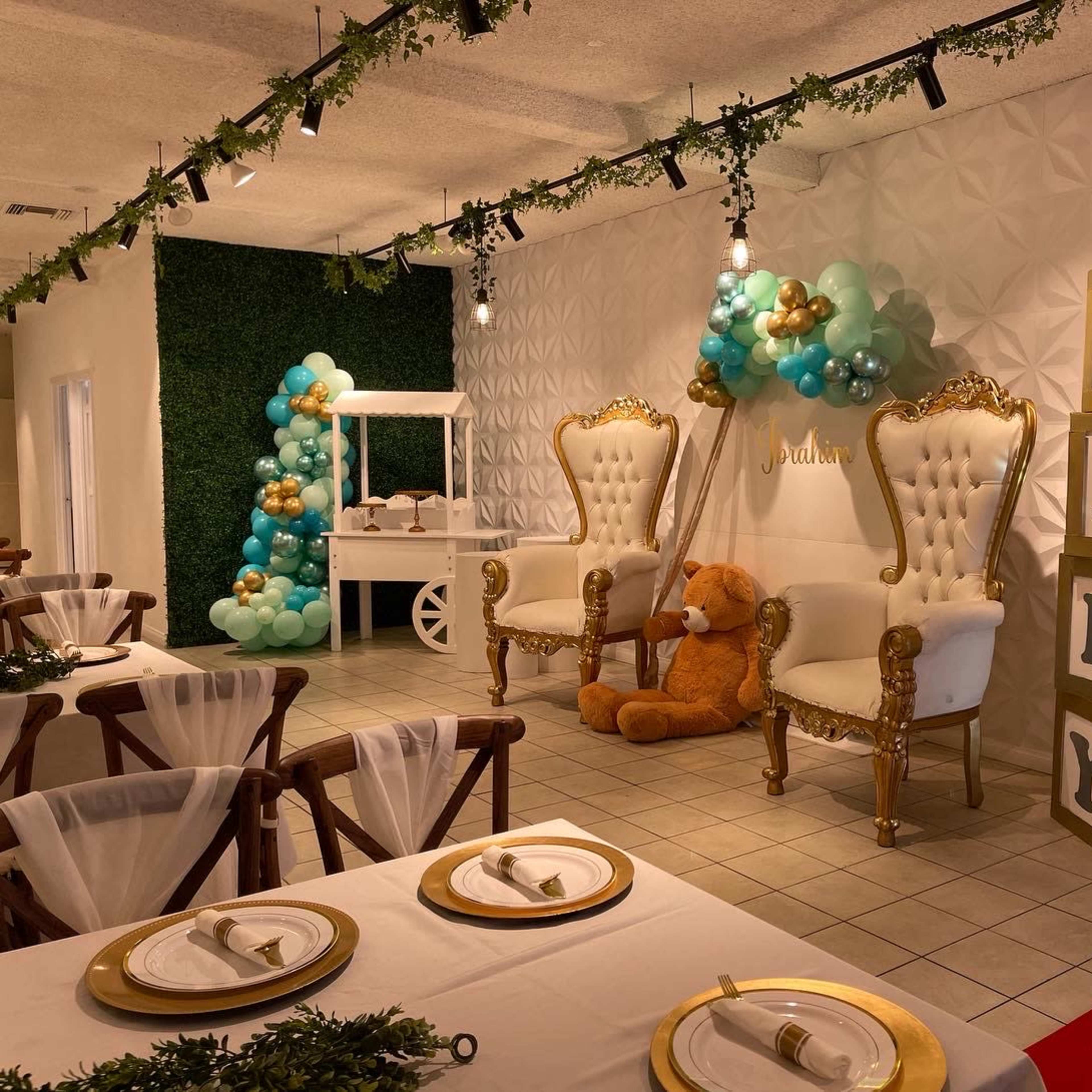 A decorated event space featuring two ornate chairs, a teddy bear, balloon arrangements, and a display of large blocks spelling "BABY."
