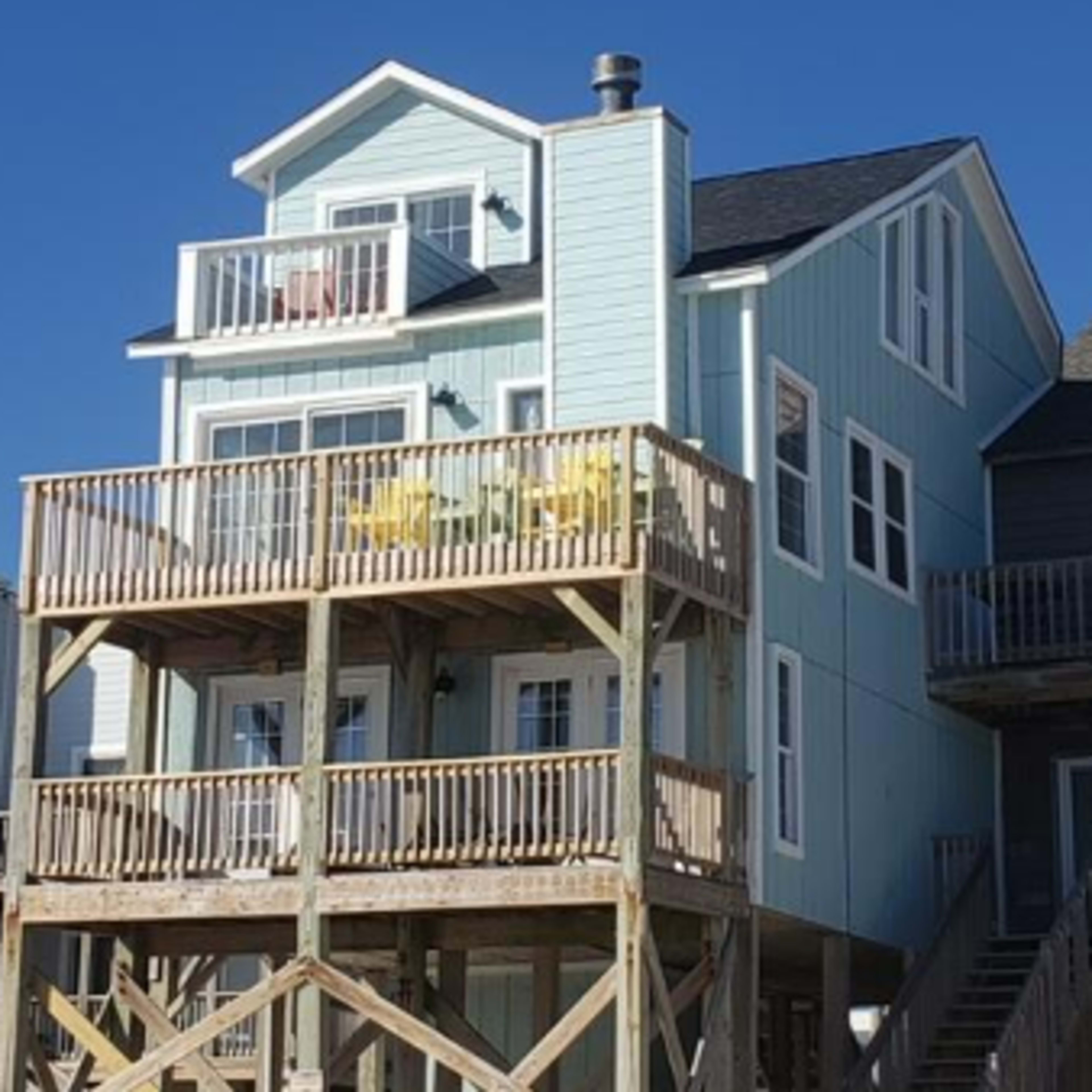 A two-story beach house with a light blue exterior and a wooden deck features two yellow chairs on the upper balcony.