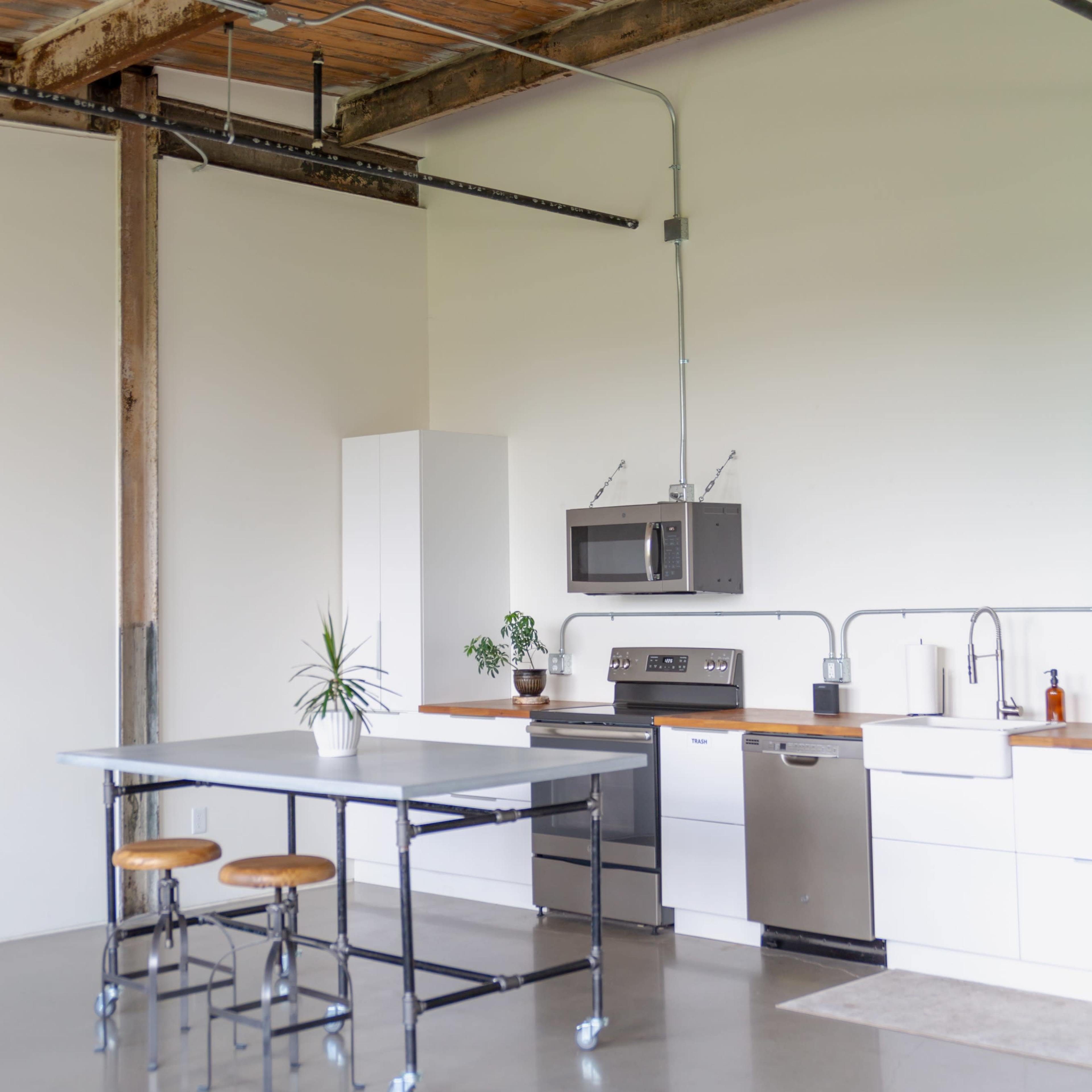 A minimalist kitchen features a central table on wheels, stainless steel appliances, and a few potted plants.