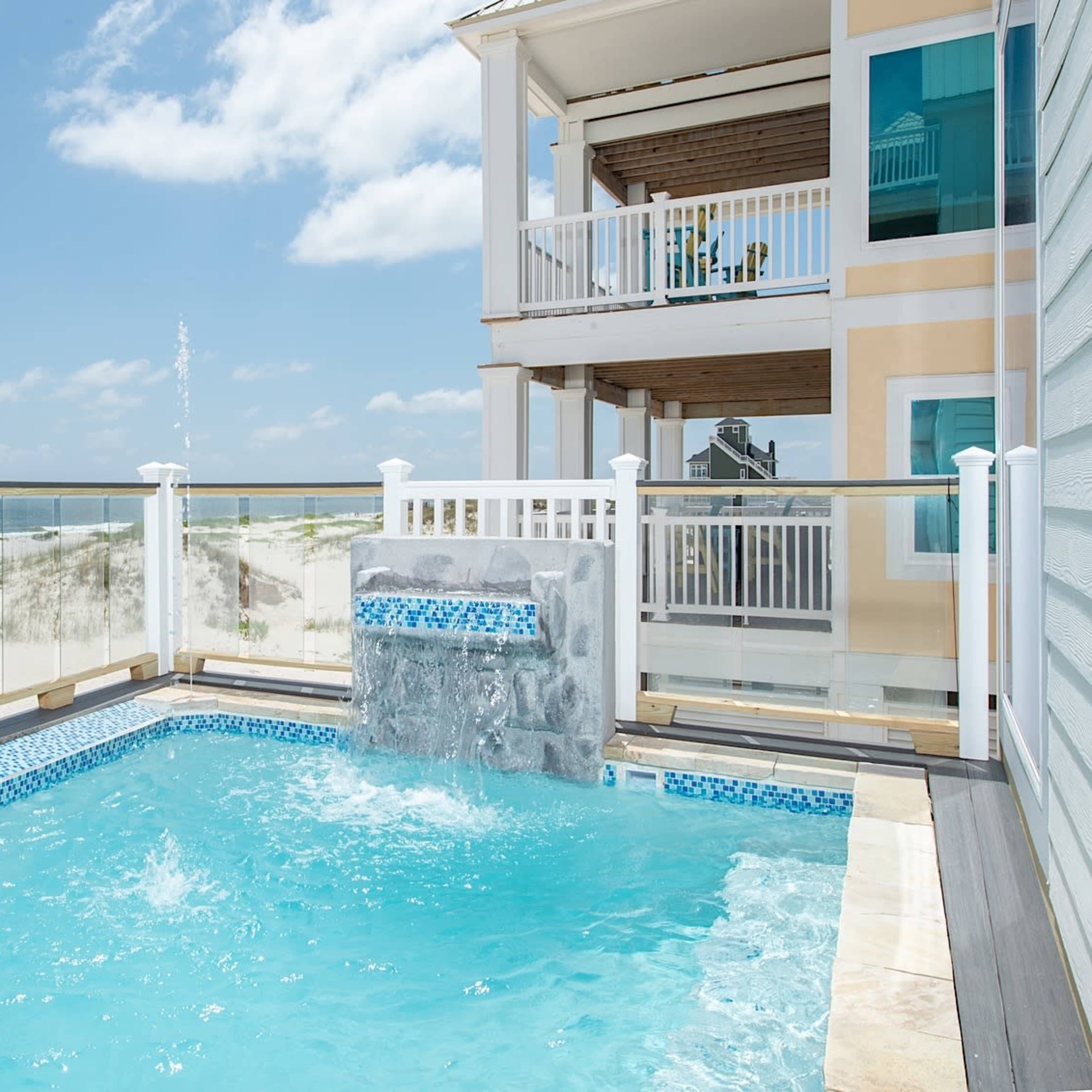 A small pool with a water feature located next to a beach house, overlooking a sandy beach and ocean.