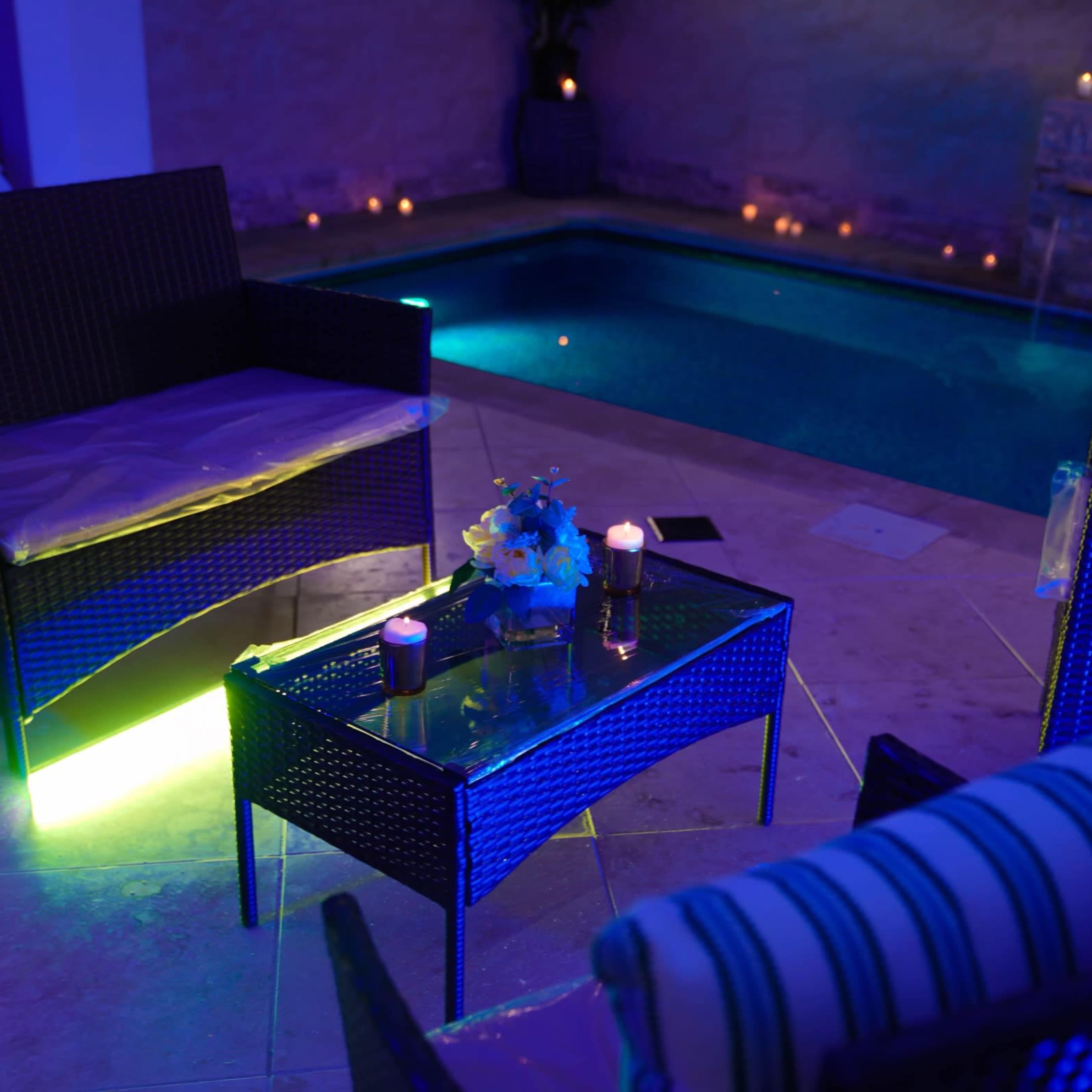 A serene poolside setting featuring two wicker chairs and a glass-top table adorned with candles and flowers, illuminated by blue lighting.