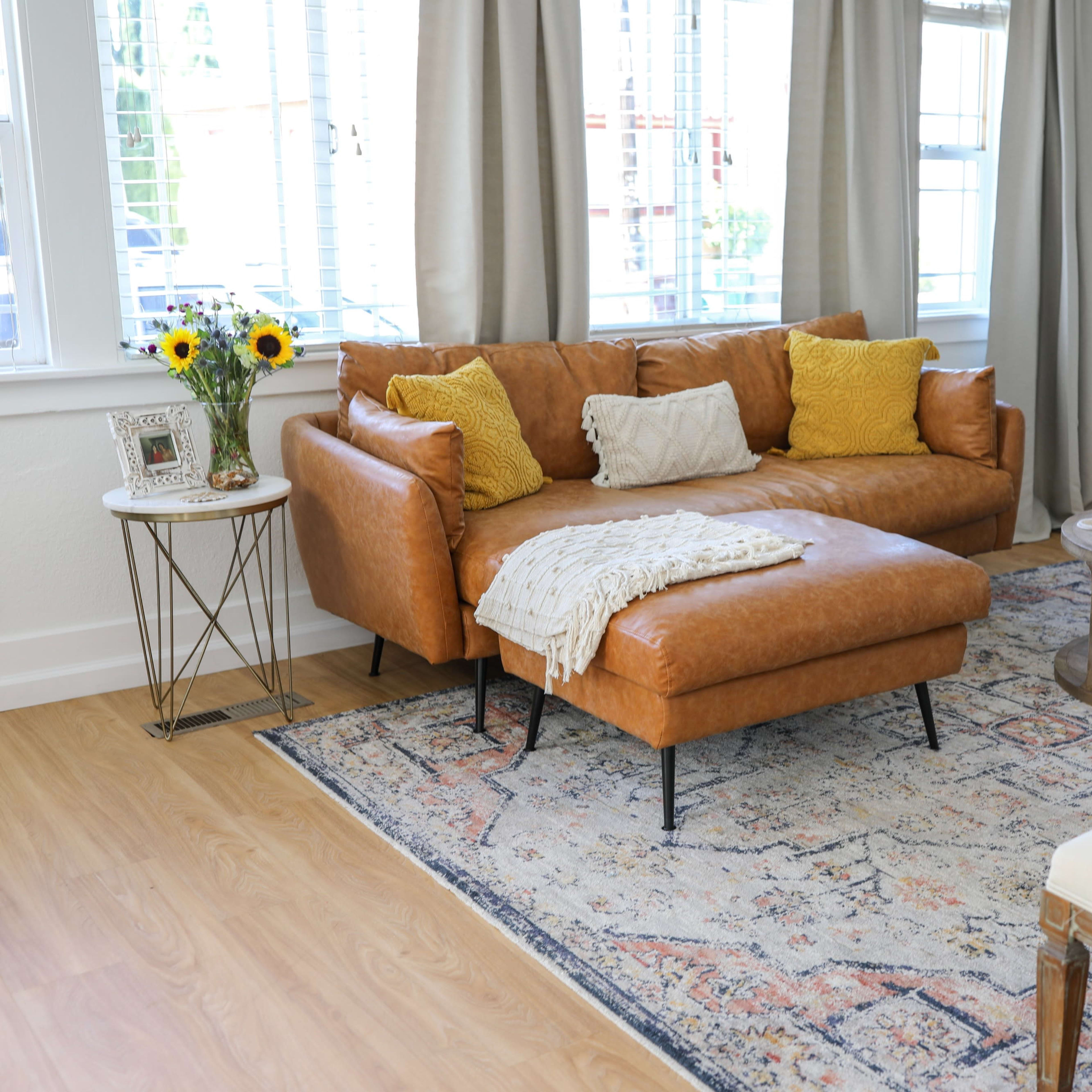 A modern living room features a brown leather sectional sofa with yellow pillows, a circular coffee table, a patterned area rug, and large windows with sheer curtains.