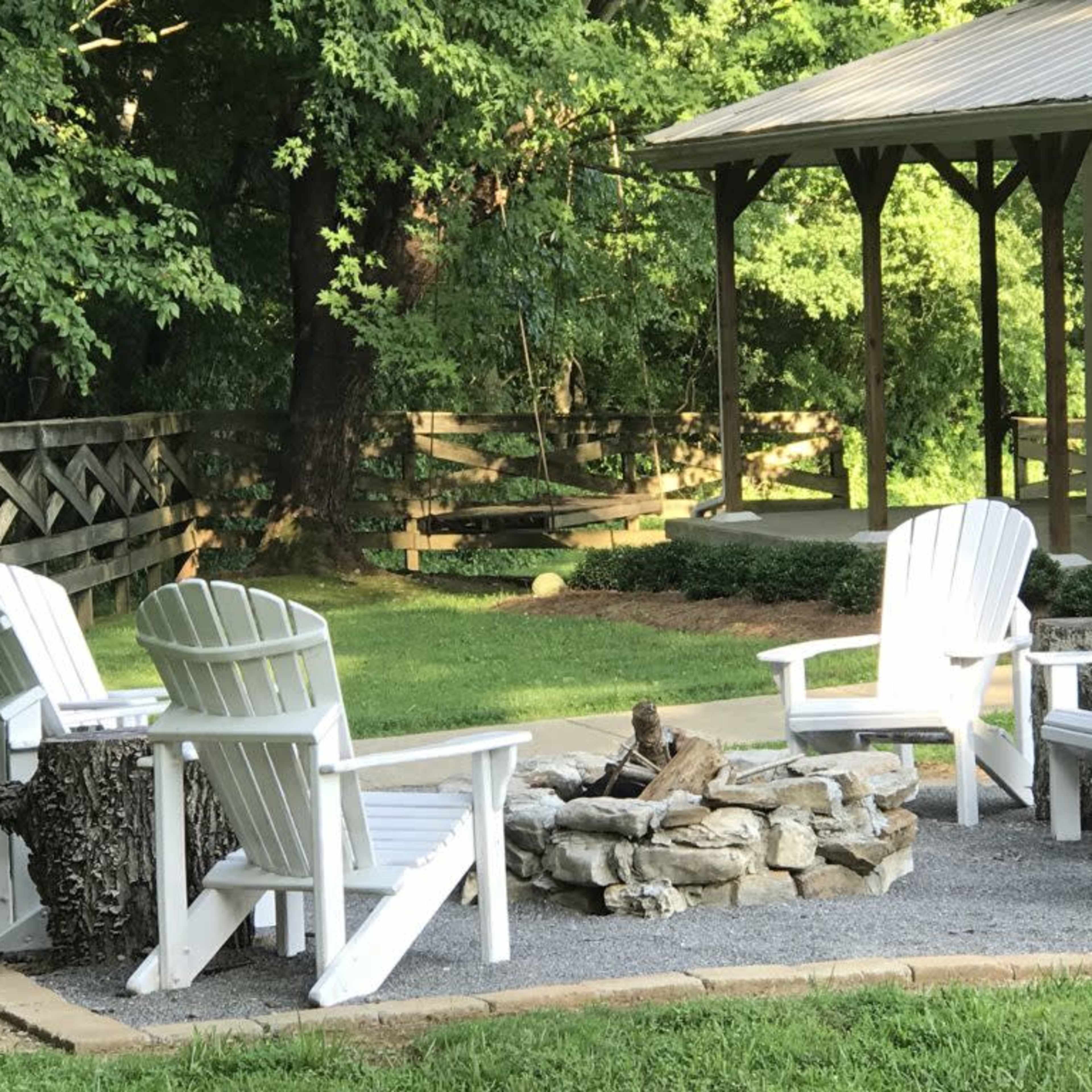 A circular seating area with white Adirondack chairs surrounds a stone fire pit, set in a grassy outdoor space near a wooden building.