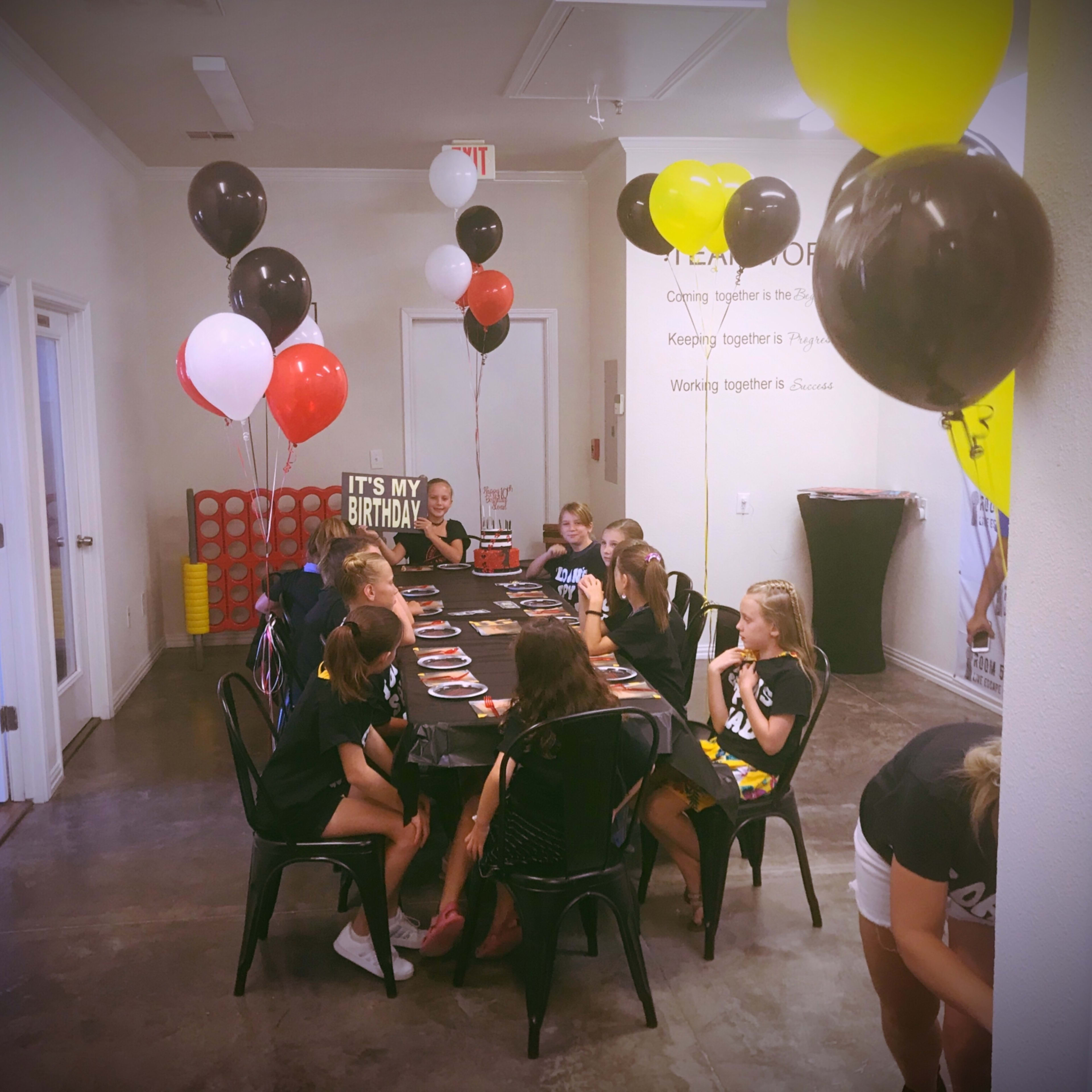A group of children is seated at a long table decorated with black and yellow balloons, celebrating a birthday party.